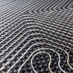 A dense grid of radiant floor heating pipes laid out on a black surface, ready for installation. The white pipes form a pattern of loops and curves over the grid, designed to distribute heat evenly when the system is in use. Some pipes have red markers.