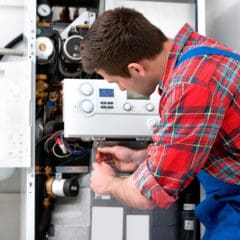 A technician in a red plaid shirt and blue overalls adjusts a component inside an open boiler system. He is focused on his task, using a screwdriver with various pipes and gauges visible around him. The setting appears to be a maintenance or repair scenario.