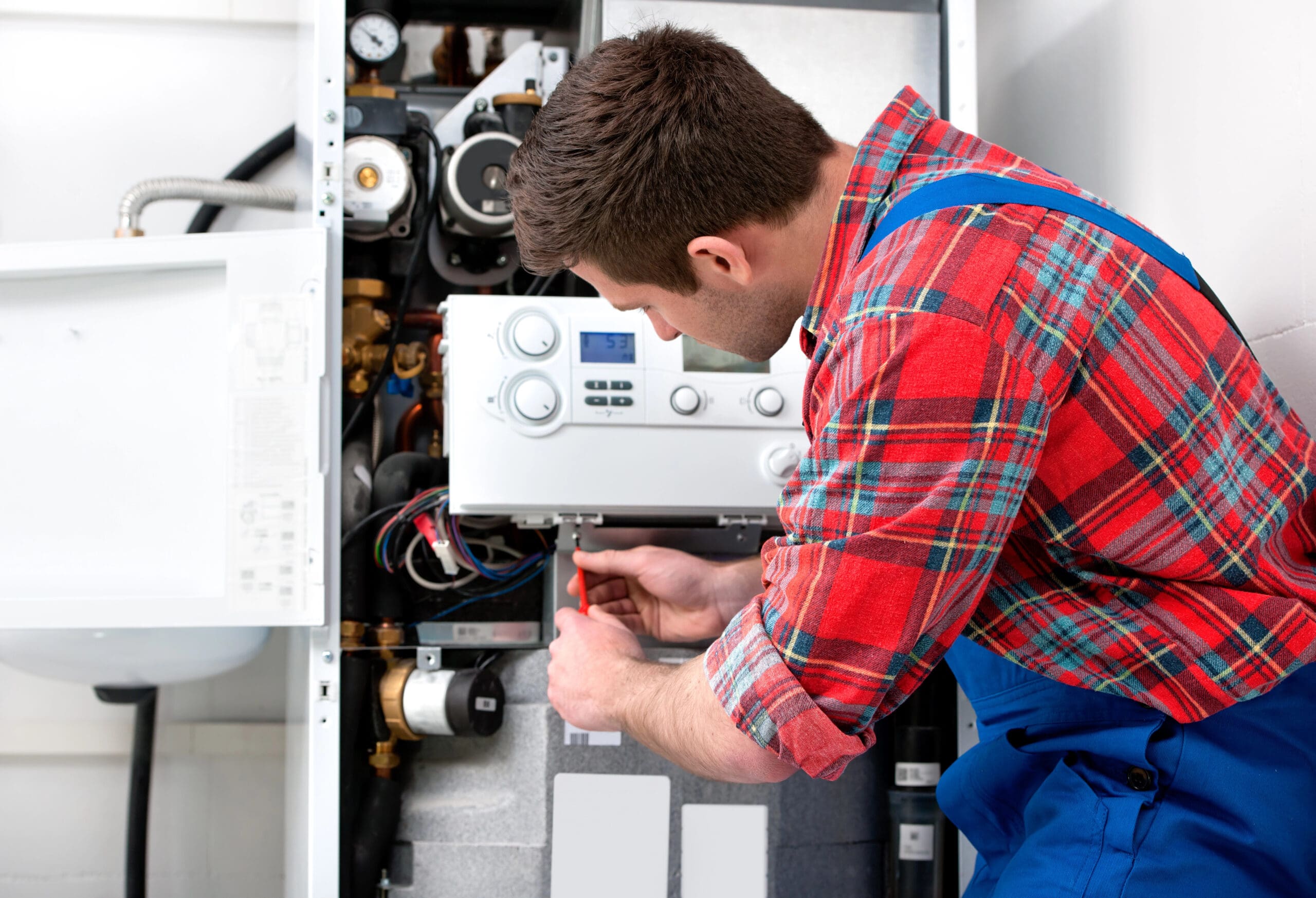 A technician in a red plaid shirt and blue overalls adjusts a component inside an open boiler system. He is focused on his task, using a screwdriver with various pipes and gauges visible around him. The setting appears to be a maintenance or repair scenario.