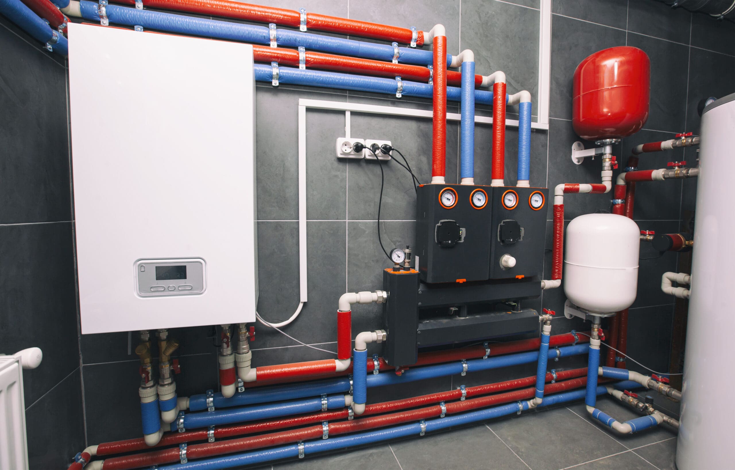 A modern boiler room with a white control unit mounted on the wall, connected to an array of blue and red pipes, gauges, and valves. The system includes a red expansion tank on the right and a series of black control units with pressure dials.