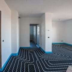 A room under construction with walls and ceiling painted white. The floor is unfinished and shows an intricate network of underfloor heating pipes, covered with a black grid structure. The room has windows and open doorways leading to other areas.