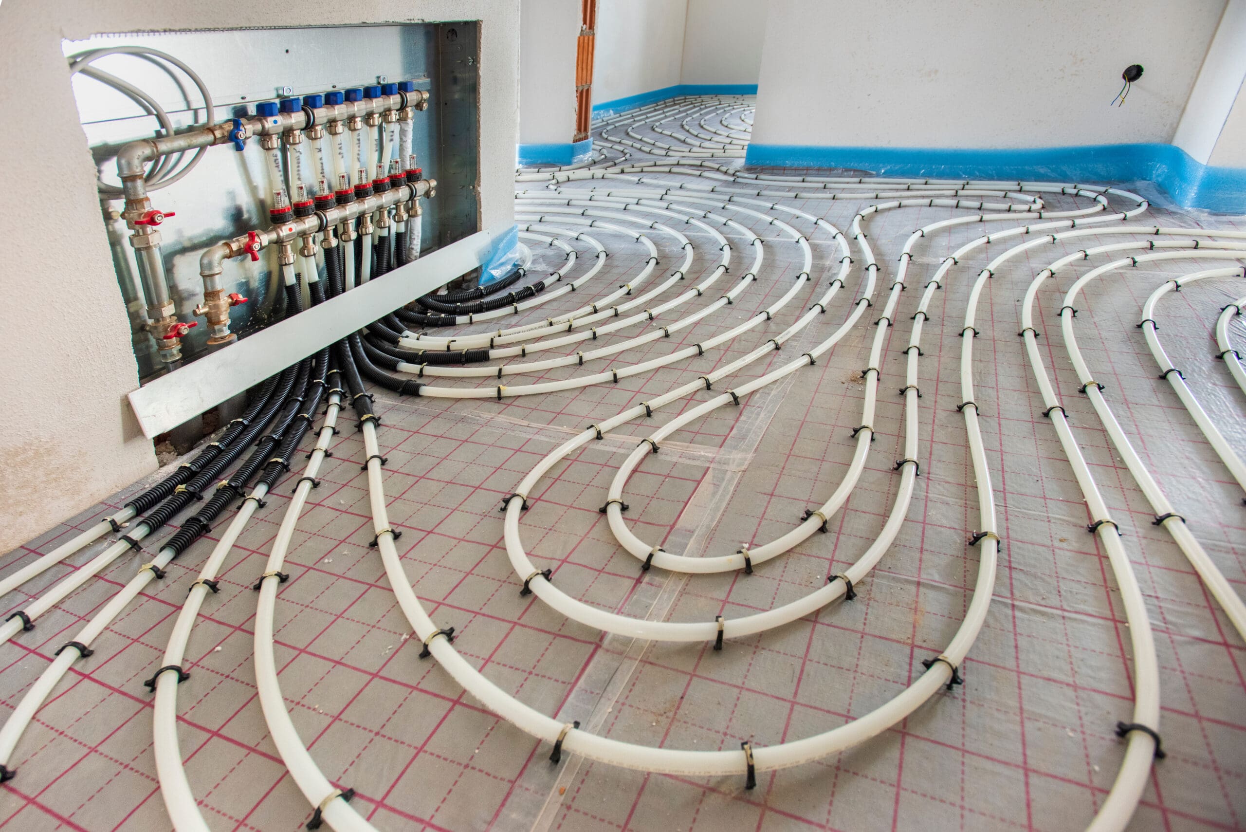 The image shows a partially installed underfloor heating system. White pipes are arranged in a serpentine pattern on the floor, connected to a manifold with multiple valves and controls on one wall. The flooring is covered with a grid-patterned insulation layer.