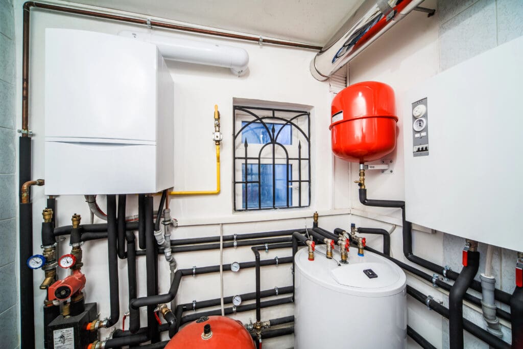 The image shows a modern boiler room with various heating equipment installed. There are white and red tanks, pipes with multiple fittings, gauges, and control panels. A small window with decorative bars is visible in the background.