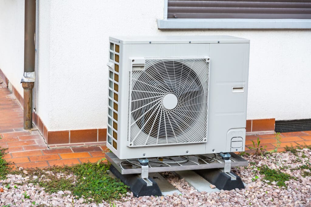 A white, outdoor air conditioning unit is mounted on a metal stand adjacent to a building with a light-colored wall. The area around the unit is covered in gravel and has some small plants growing. A brown downspout is attached to the building wall.