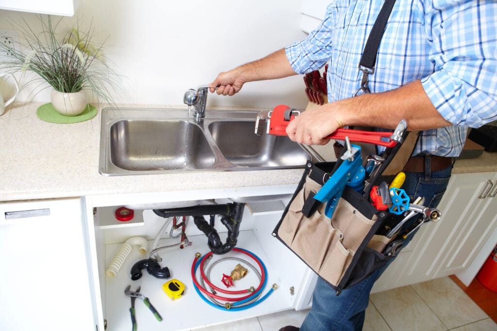 A person wearing a blue plaid shirt and carrying a tool bag is working on a kitchen sink. Various tools are visible in the tool bag and on the counter. The cabinet under the sink is open, exposing plumbing pipes and additional tools.