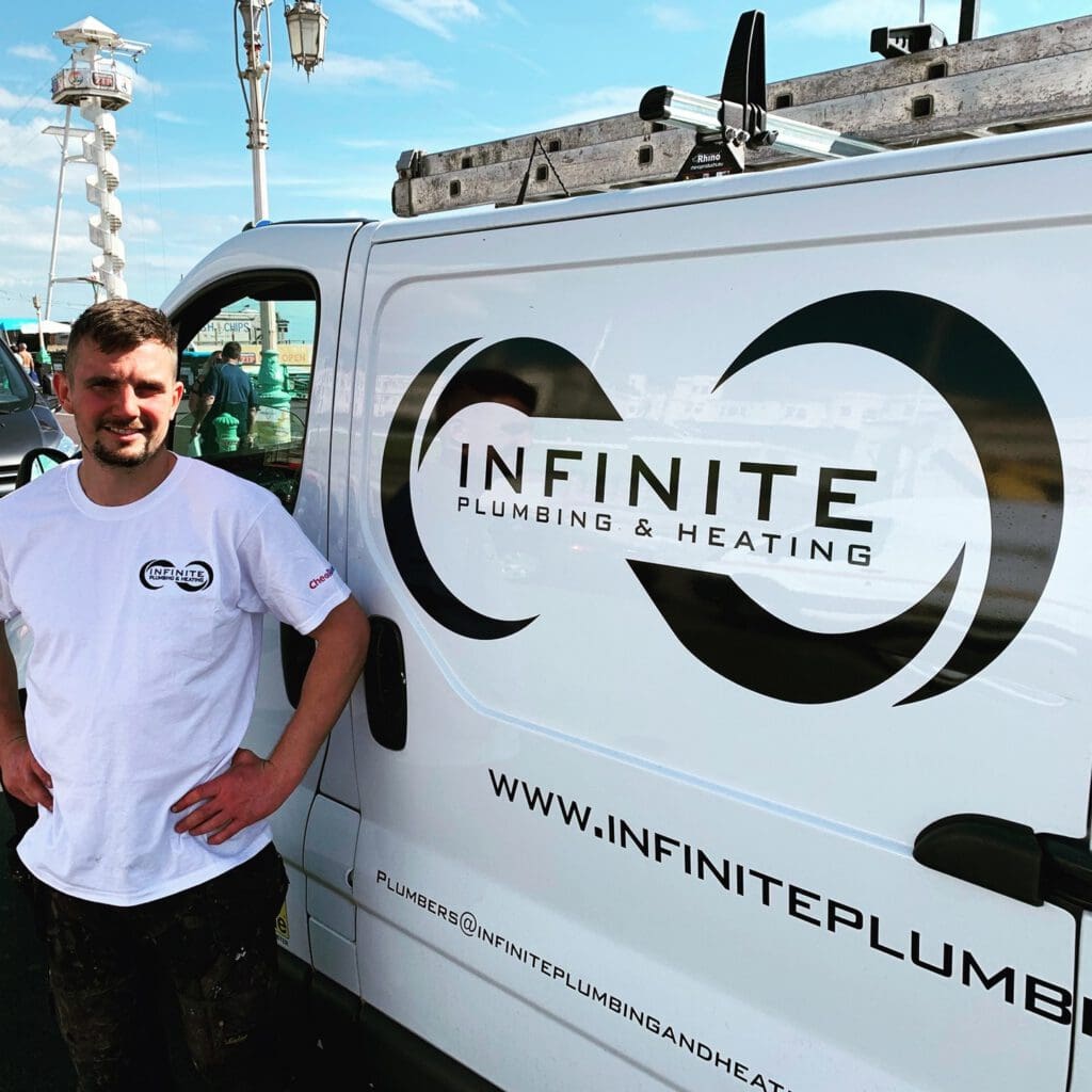 A man wearing a white t-shirt stands beside a van with the logo "Infinite Plumbing & Heating." The van’s logo features an infinity symbol, and the website and contact information are displayed on the side of the van. A lighthouse is visible in the background.