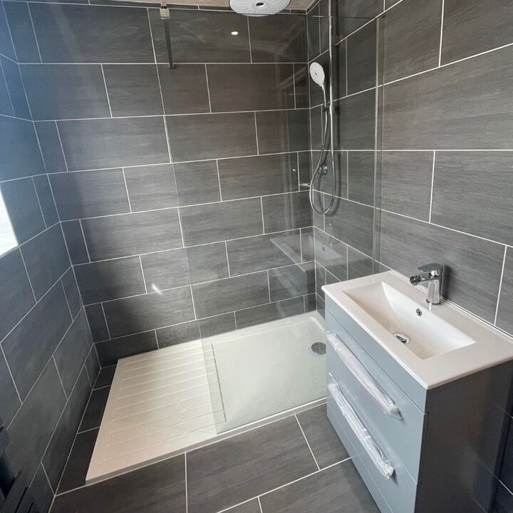 A modern grey-tiled bathroom features a sleek walk-in shower with a glass partition and a white shower tray. An overhead rain shower and a handheld showerhead are visible. Next to the shower is a grey vanity unit with a white sink and a chrome faucet.