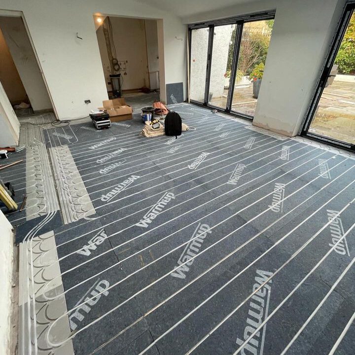 A room is under renovation with large sliding glass doors. The floor is covered with Warmup heating mats, indicating the installation of underfloor heating. A person works kneeling on the floor amidst tools and boxes, preparing the space for further construction.