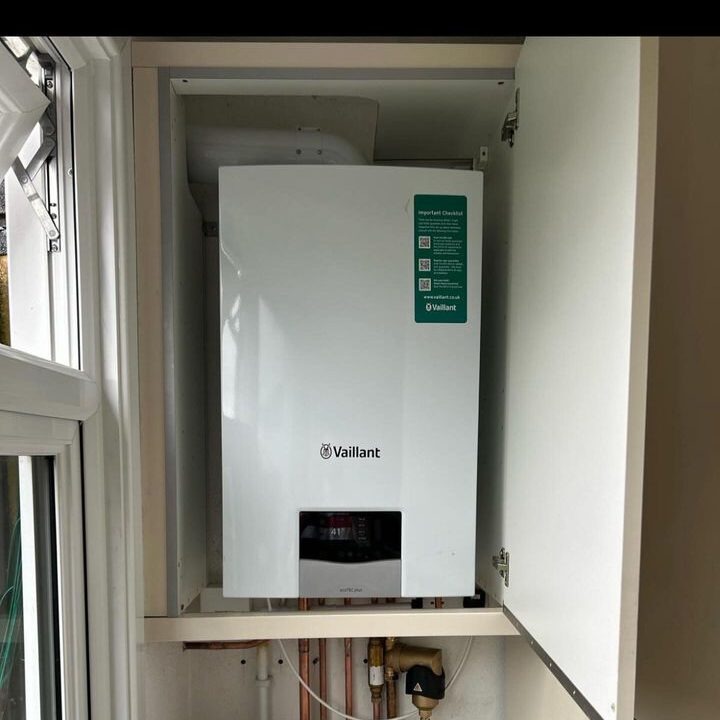 A white Vaillant boiler is installed inside a compact, open cabinet. The boiler features a small, rectangular display panel and has pipes and electrical connections visible below it. The cabinet has open doors and is situated next to a window.