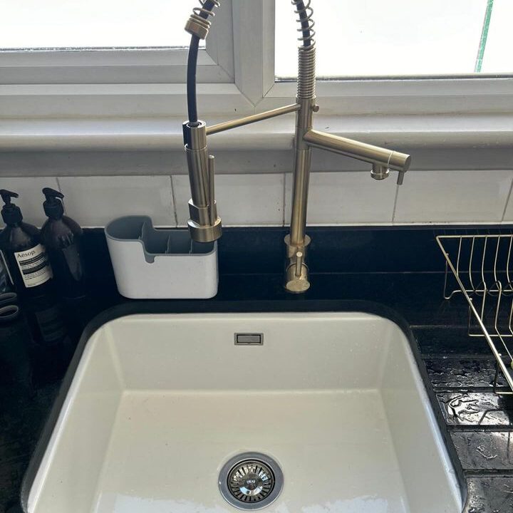 A clean white kitchen sink with a modern brass faucet under a window. To the left of the faucet, there is a white utensil holder attached to the backsplash with some items inside. On the right, part of a dish rack is visible.