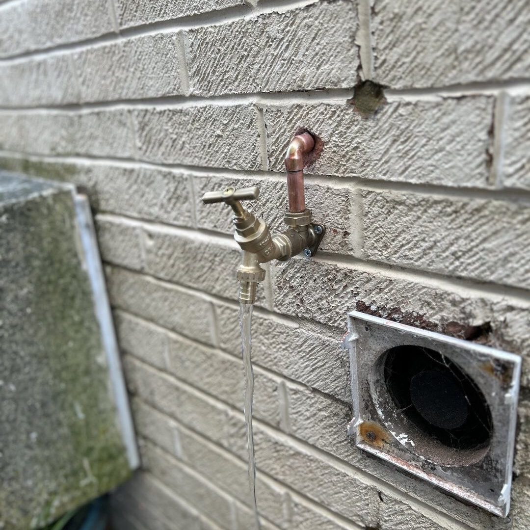 An outdoor water spigot is mounted on a brick wall, with a hose attached. The spigot is connected to copper piping. Below the spigot, there is a vent with a cracked frame. The brick wall has some wear and tear. Green and gray structures are partially visible to the left.