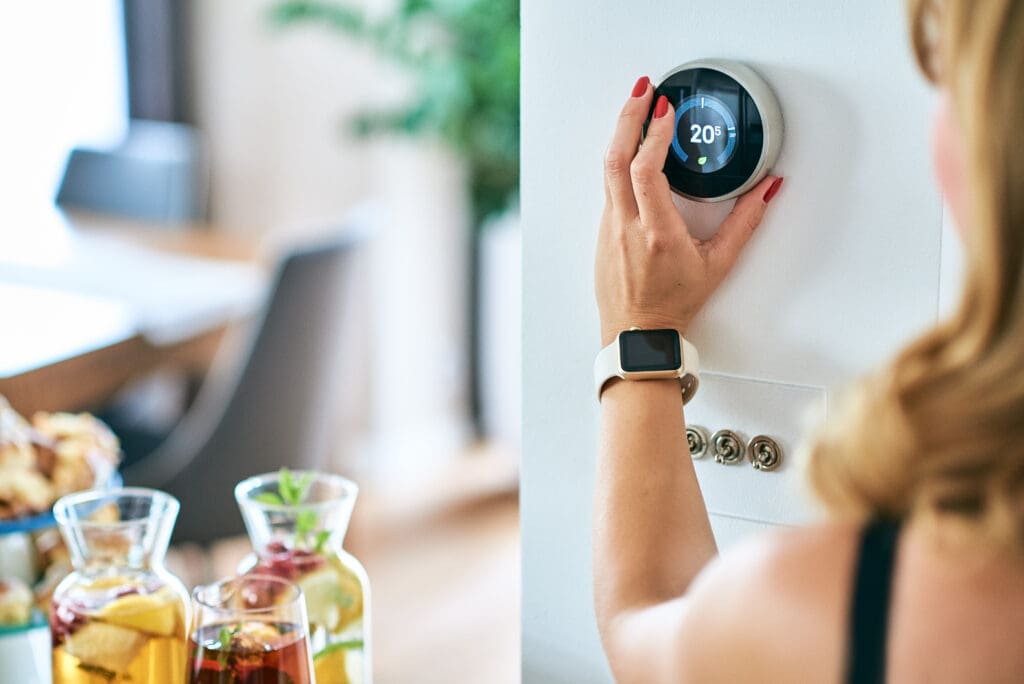 Person adjusting a modern thermostat on a wall. The thermostat displays "20°." The person is wearing a smartwatch. A dining table with glasses of iced beverages and a plate of food is visible in the background. The setting appears to be a comfortable home.