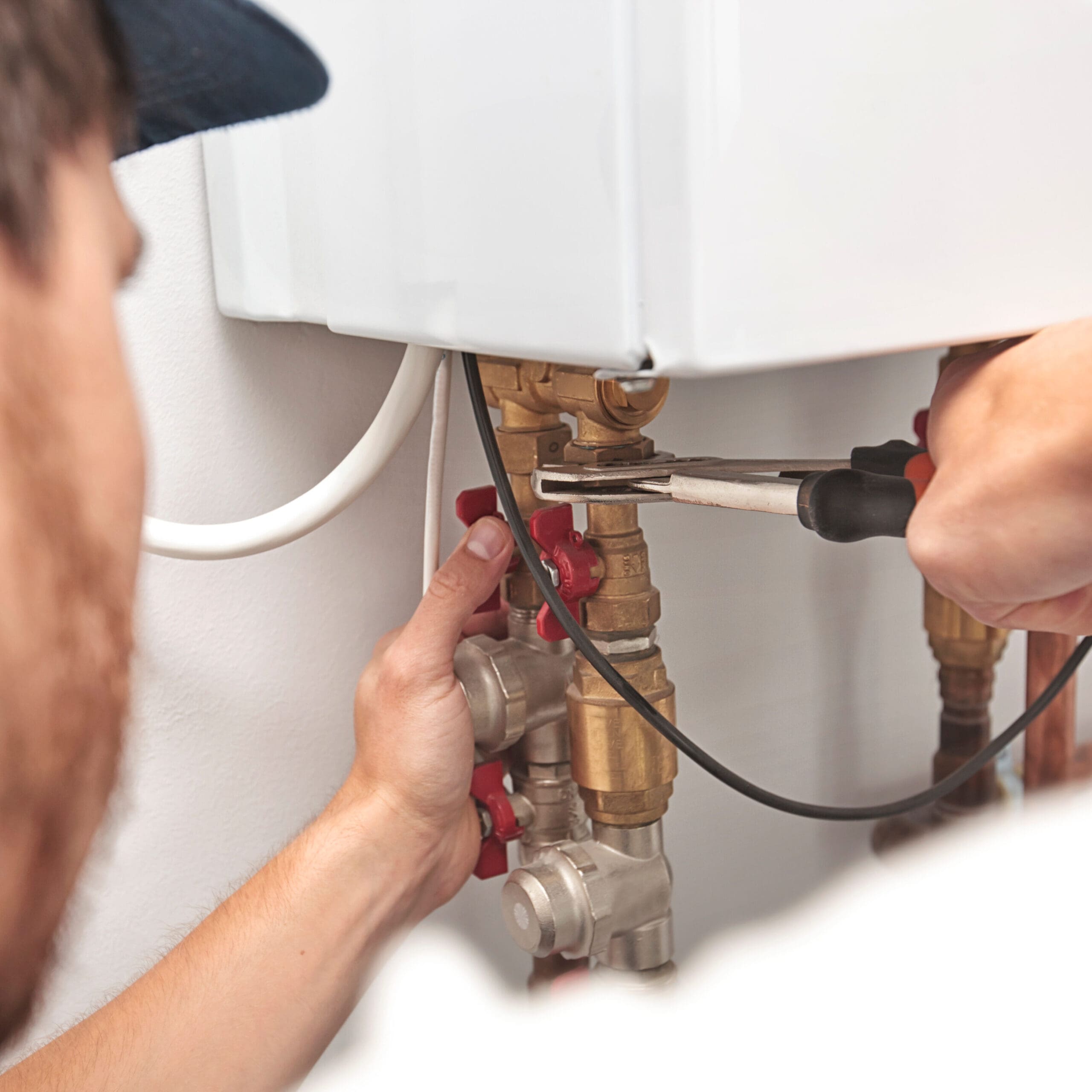 A plumber using a wrench to tighten a valve under a white boiler. The plumber is partially visible, wearing a blue cap. Copper and brass pipes with red knobs are shown, and a white electrical cable is visible. The focus is on the plumber's hands and tools.