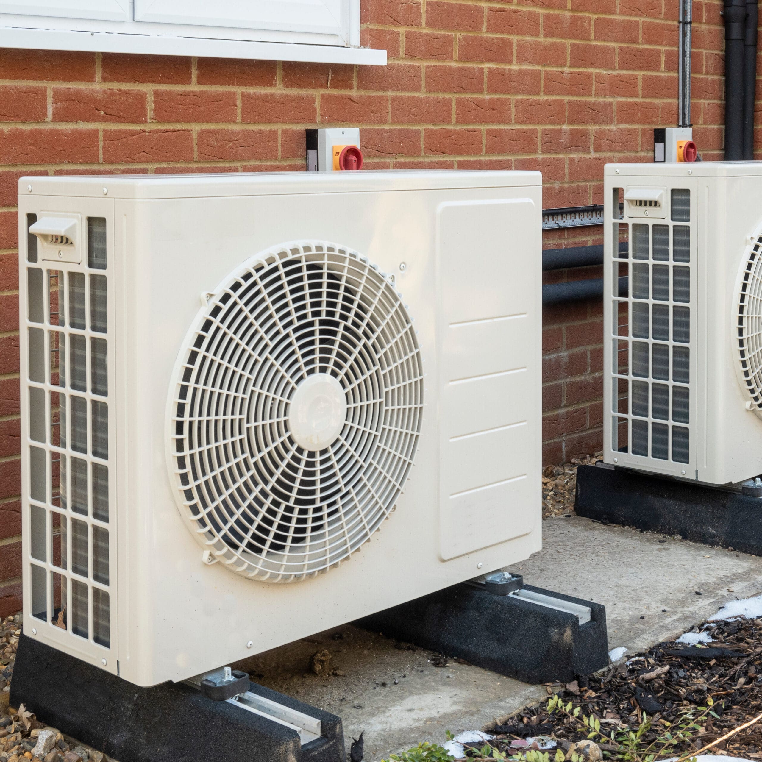 Two outdoor air conditioning units are installed against the brick wall of a building. Each unit is mounted on a raised platform and has a large, white fan grille on the front. The area around the units is paved and has gravel.