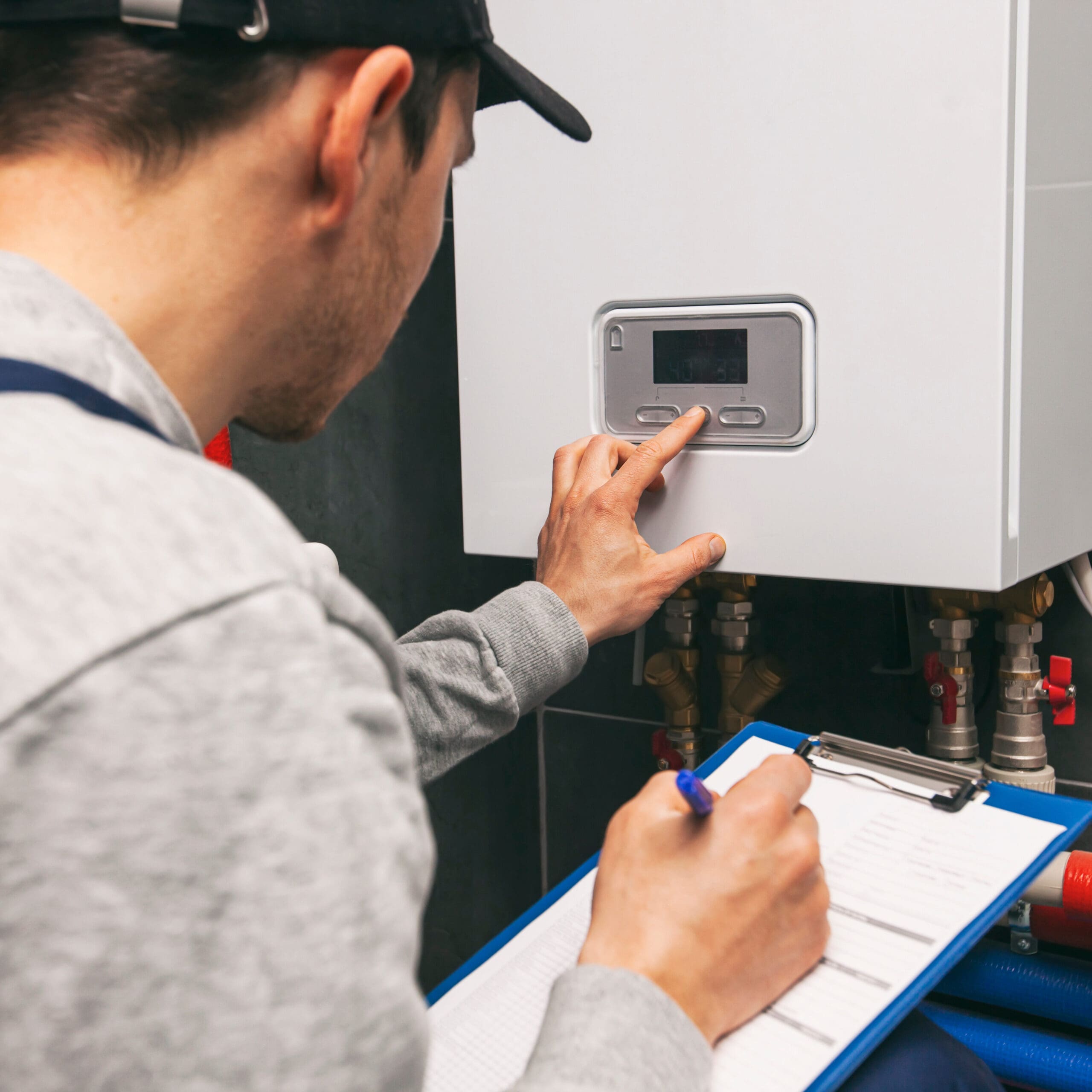 A technician in a gray sweatshirt and black cap is facing away from the camera, operating a control panel on a wall-mounted device. He is pressing buttons while holding a blue clipboard and pen, likely noting down readings or making adjustments.