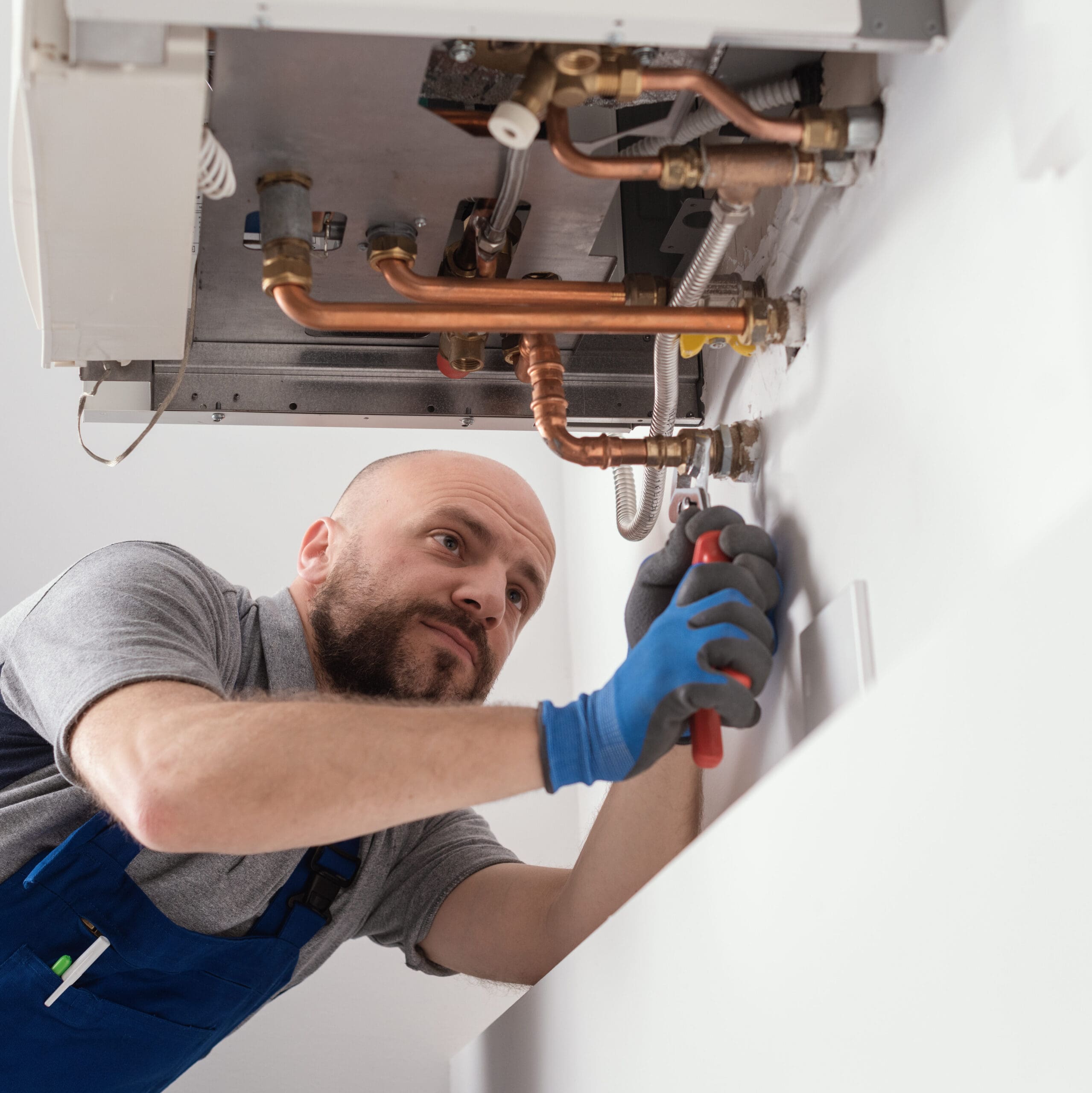 A repairman with a beard is working with tools on a boiler or heating system mounted on a wall. He is wearing a grey shirt, blue overalls, and blue gloves, and he appears focused on his task. Copper pipes and various fittings are visible above him.