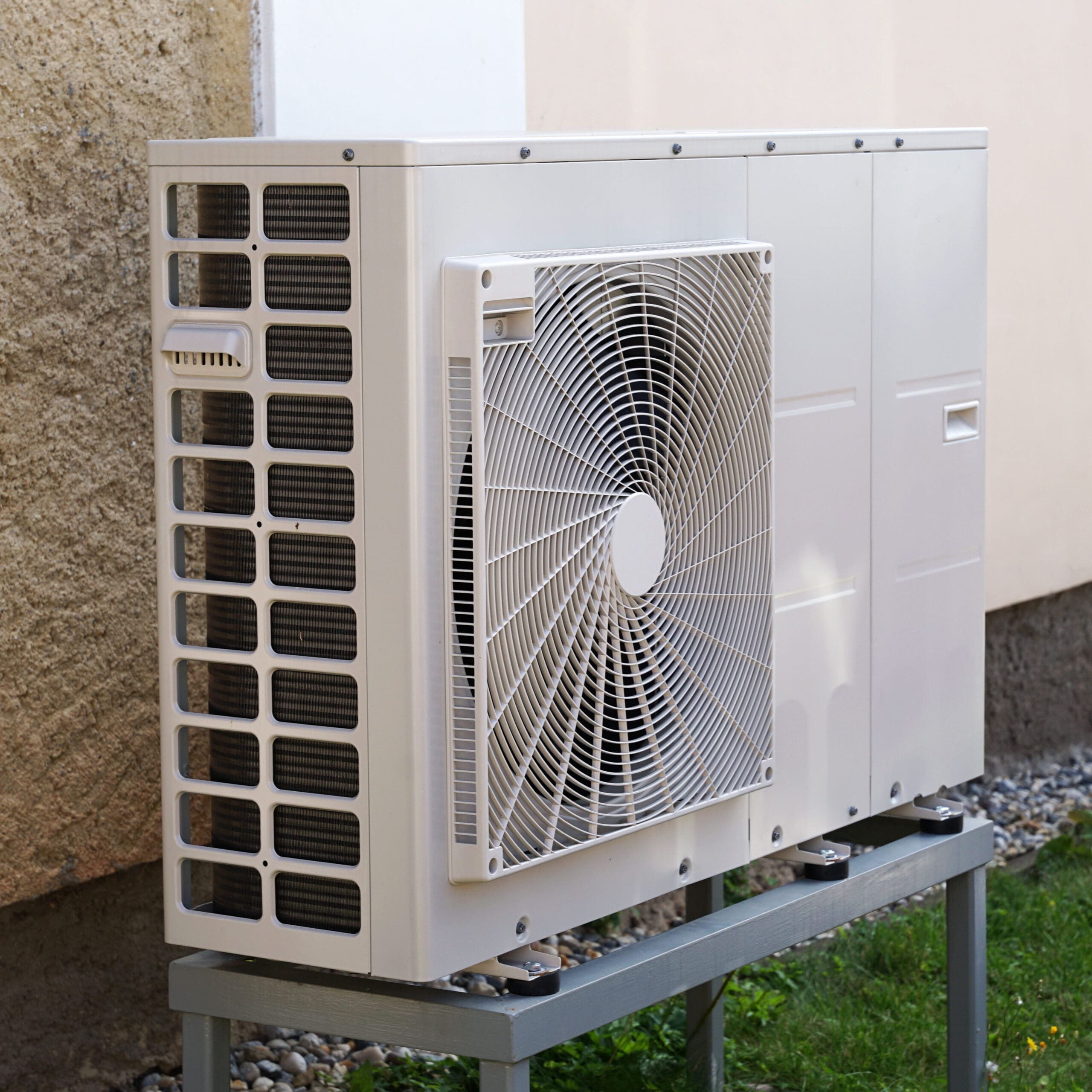 Image of an outdoor heat pump unit mounted on a metal stand against the exterior wall of a building. The unit features a large fan in the center and multiple air vents on the sides.