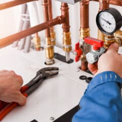 A person in a blue jacket is adjusting a valve on a network of copper pipes connected to a gauge. The person is holding a wrench in their left hand, tightening a fitting. The setup appears to be part of a heating or plumbing system.