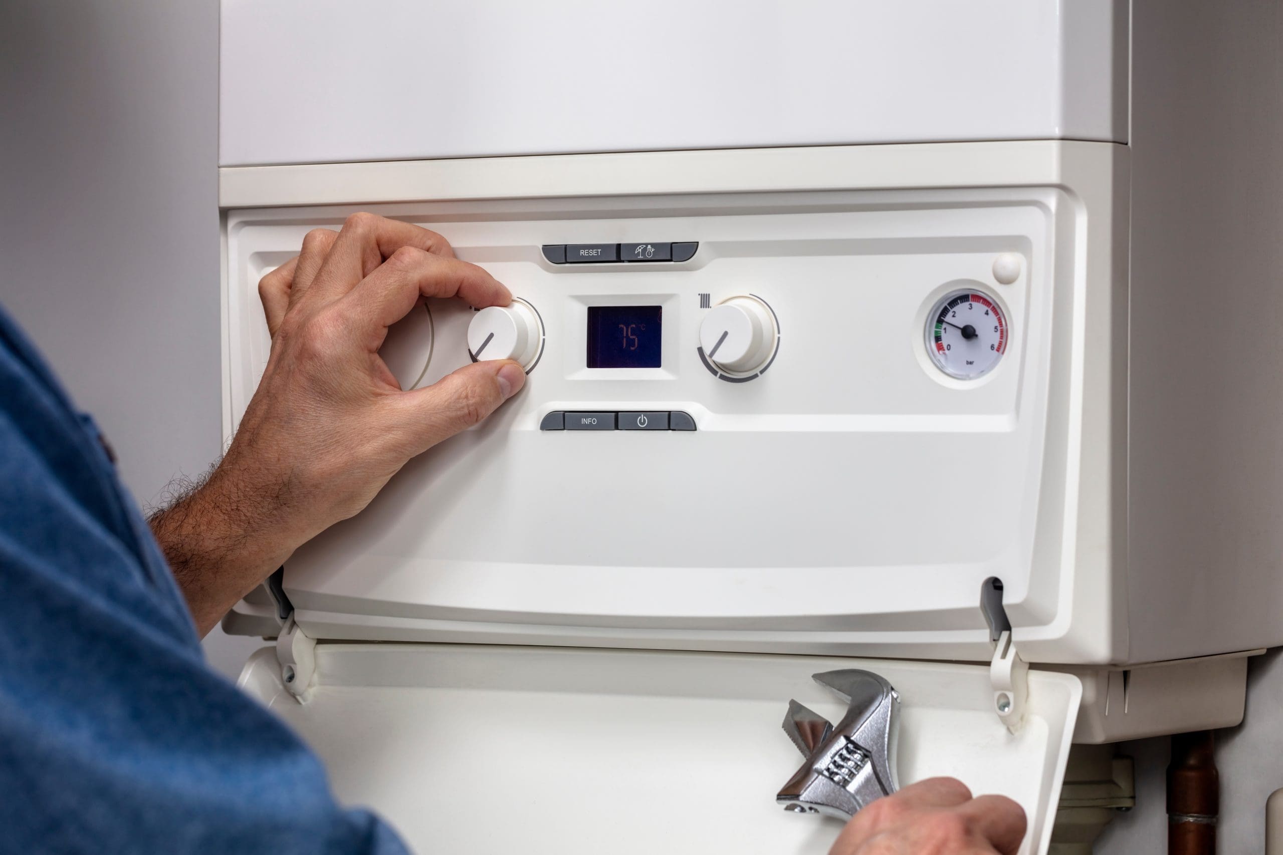 A person adjusts the settings on a white home heating boiler, turning a dial with their left hand. Their right hand holds an adjustable wrench resting on the boiler. The boiler's digital display reads "75," and a pressure gauge is visible on the right.