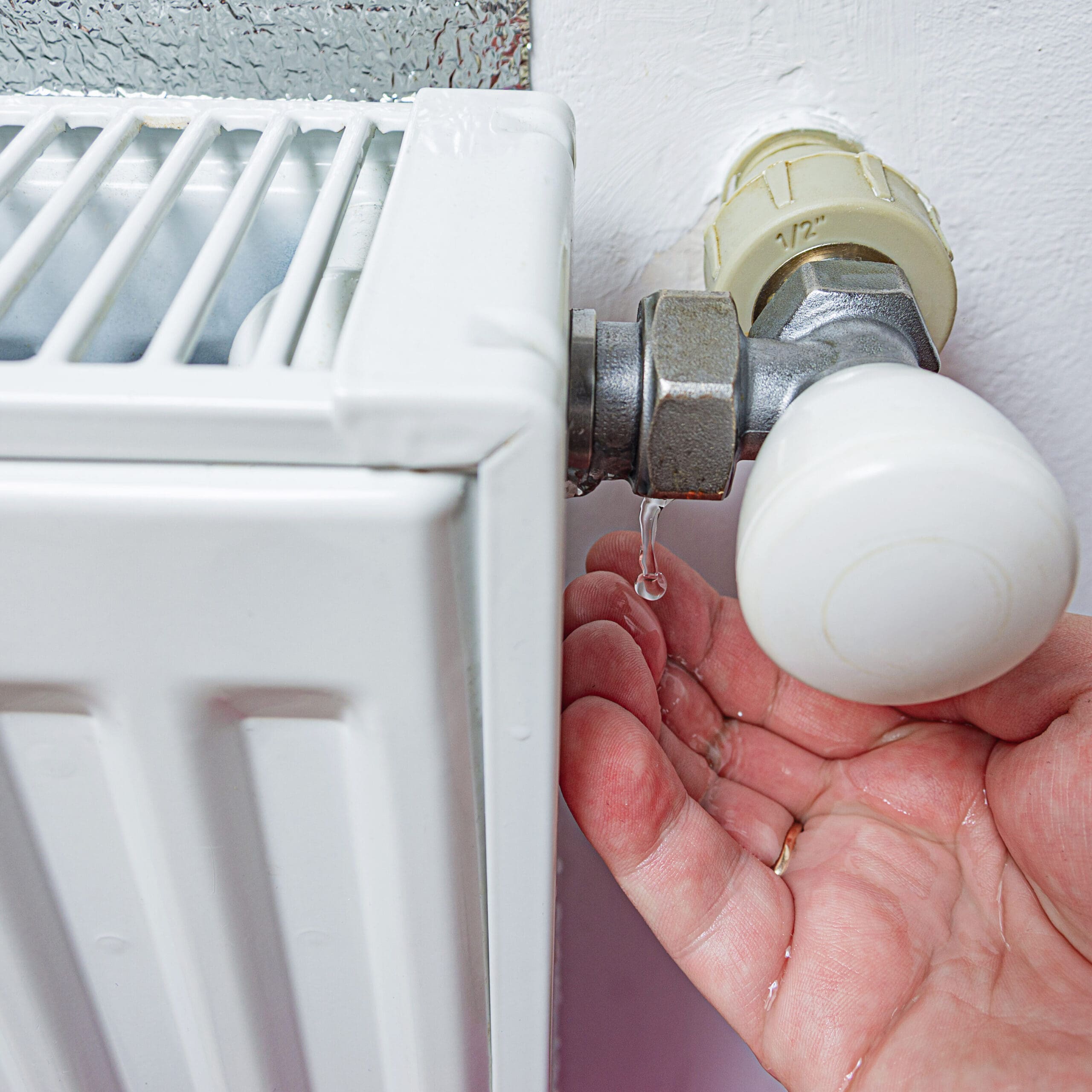 Close-up of a hand collecting a droplet of water from a white radiator valve. The radiator and valve are attached to a white wall. The hand is under the valve, indicating a leak. The image focuses on the leakage issue.