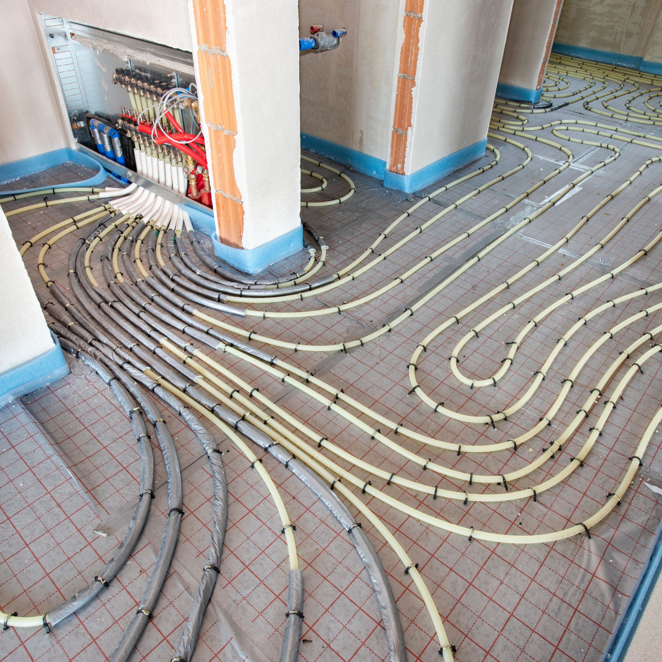 A room under construction with exposed underfloor heating pipes arranged in a serpentine pattern. The walls are not yet plastered, revealing their concrete and brick structure. Multiple pipes are connected to a control panel embedded in a wall.