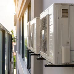 Two white outdoor HVAC units mounted on the exterior wall of a modern building. The units are installed one above the other on metal brackets. The glass balcony railing and the building's windows are visible in the background, with sunlight shining through.