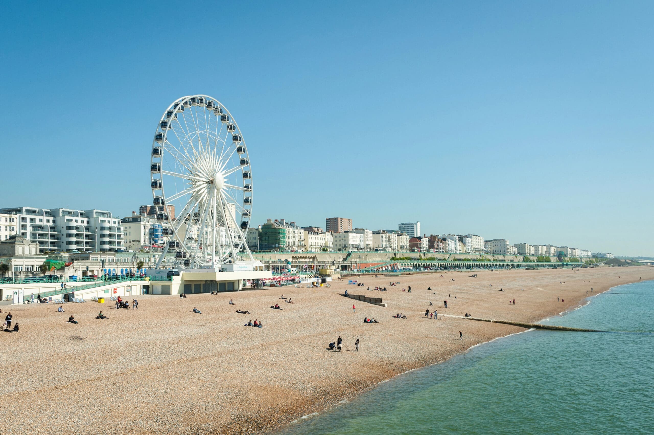 A clear, sunny day at a pebbled beachfront with people relaxing and a large Ferris wheel as a focal point. The coastline is lined with buildings, and the calm sea stretches out to the horizon.