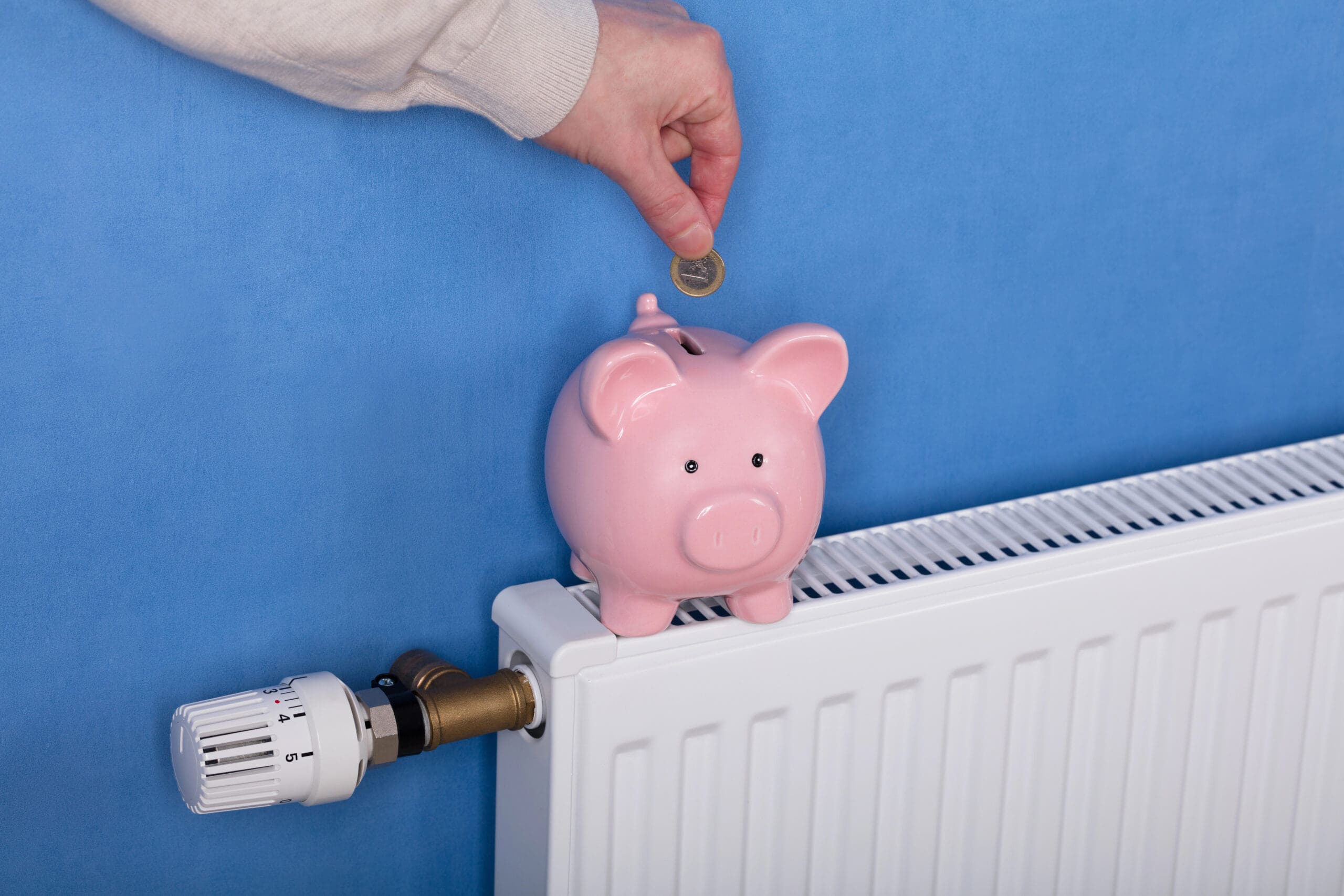 A person's hand placing a coin into a pink piggy bank sitting on a white radiator with a temperature control knob on the left, positioned against a blue wall.