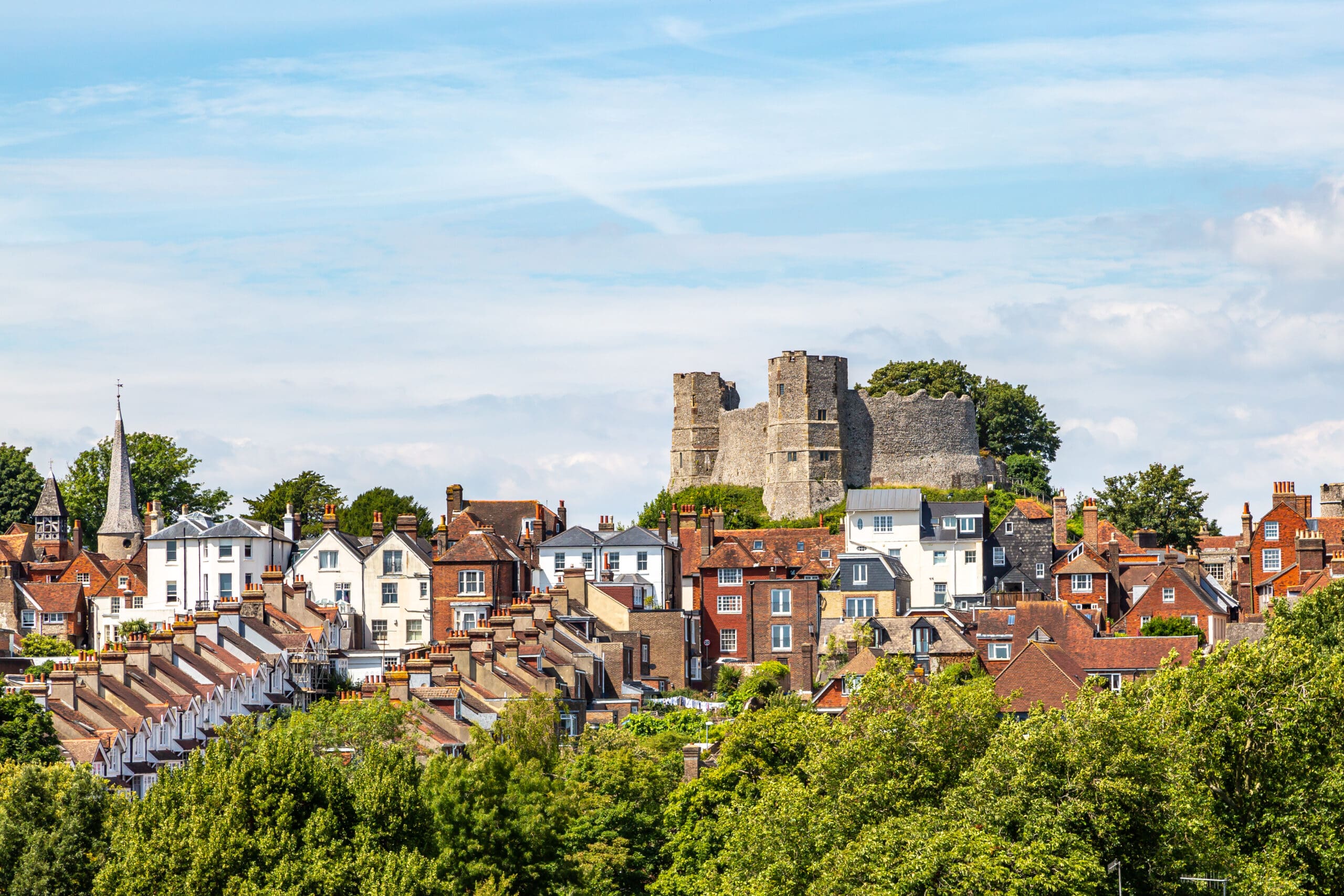 A scenic view of a historical town featuring rows of houses with red-tiled roofs and a prominent medieval castle atop a hill in the background. Lush green trees and a church steeple are also visible under a blue sky with light clouds.