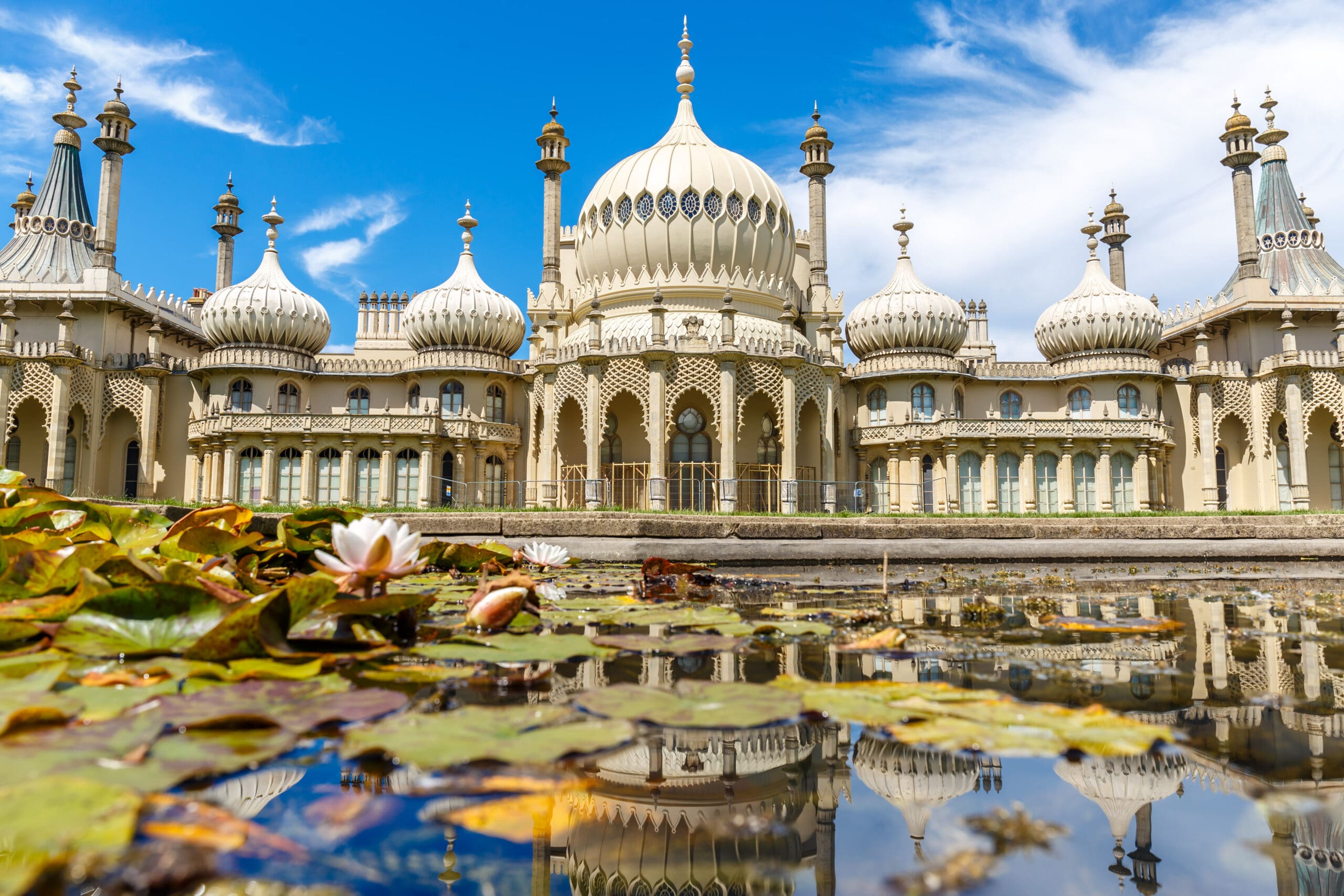 The image shows the Royal Pavilion in Brighton, UK, a historic palace with an exotic style. The architectural details include domes, minarets, and intricate designs. In the foreground is a serene pond with lily pads, reflecting the structure under a bright blue sky.