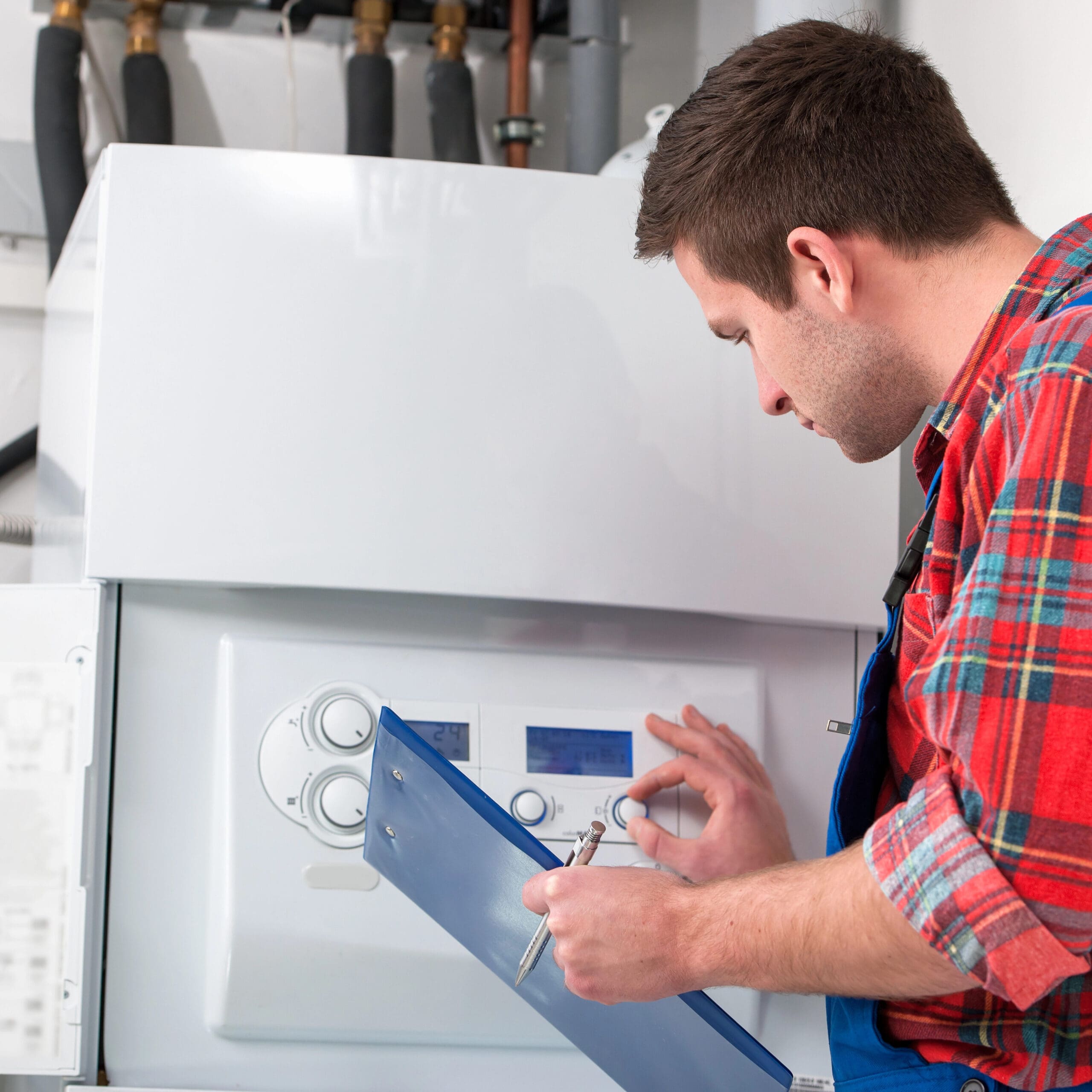 A technician in a plaid shirt and overalls is adjusting the controls on a boiler system while holding a clipboard and pen. The technician is focused, ensuring the system is properly configured. The background shows various pipes and equipment.