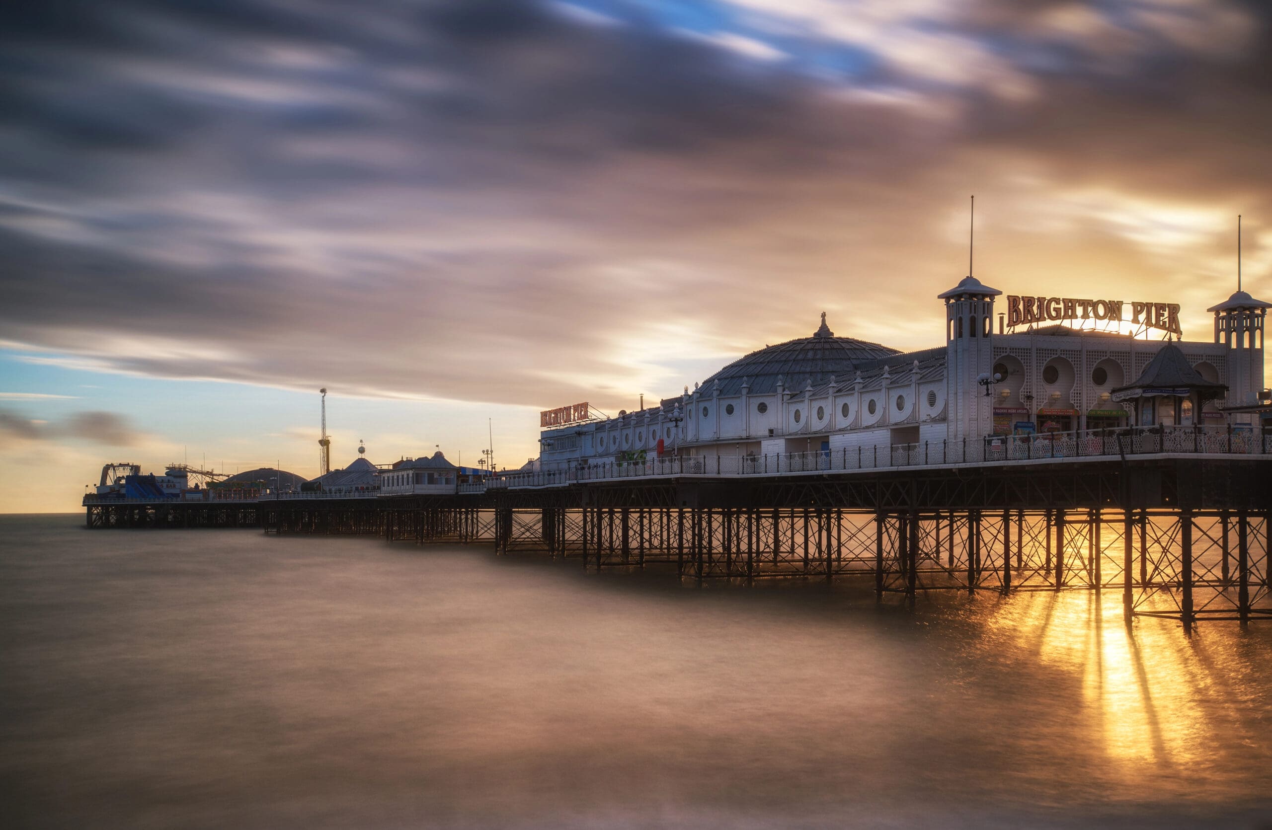 Image of Brighton Pier extending over calm sea water at sunset. The sky is filled with dramatic clouds, with rays of sunlight breaking through, casting a golden glow over the pier. The text "Brighton Pier" is visible on the structure.