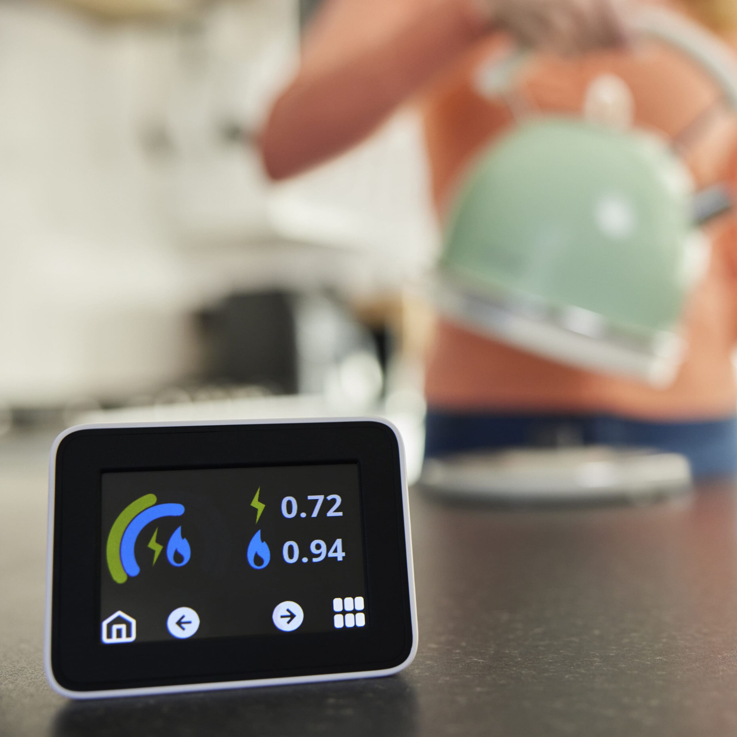 A person in the background is holding a pastel green teapot. In the foreground, there's a small digital screen displaying electricity and gas usage in units of 0.72 and 0.94 respectively. The screen is placed on a dark surface.