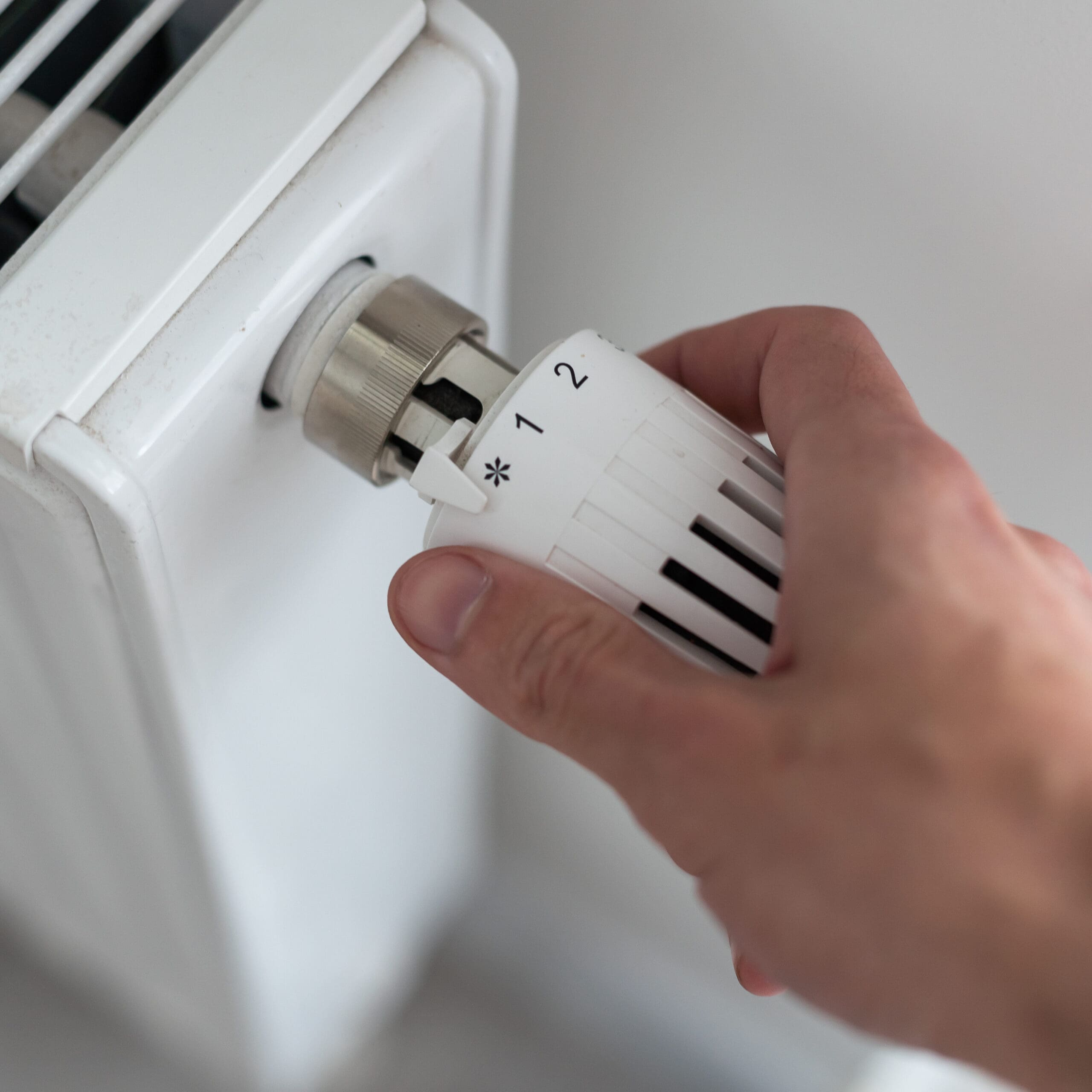 A hand turning the knob on a radiator thermostat to adjust the temperature. The thermostat displays settings labeled with a star, 1, and 2. The radiator is white with horizontal slats.