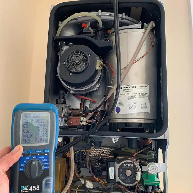 A person holds a digital combustion analyzer displaying readings in front of an open boiler system. The boiler's internal components, including a fan, pipes, and a labeled cylinder, are visible. The display shows various gas measurements and statistics.