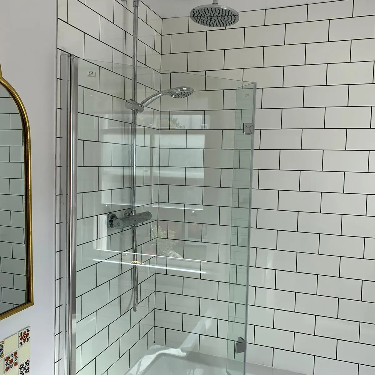 A modern bathroom with white subway tiles features a glass-enclosed corner shower. The shower has a rainfall showerhead from the ceiling and a handheld showerhead on the wall. To the left, part of a mirror with a decorative frame is visible.