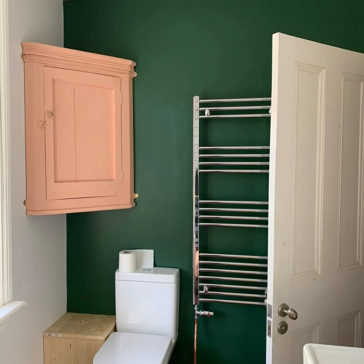 A bathroom with dark green walls features a closed peach-colored wall cabinet, a white toilet with a roll of toilet paper on top, a chrome towel rack, and a part of a white door and sink. The room has wooden flooring and a window that partly illuminates the space.