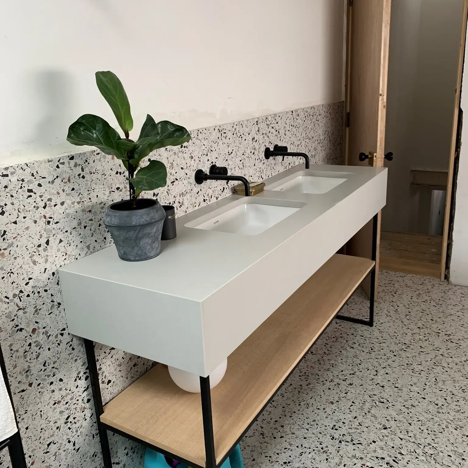 A modern double sink bathroom vanity with white rectangular basins and black fixtures stands against a terrazzo-tiled wall. A potted plant sits to the left on the white countertop, and a wooden shelf below provides additional storage. A door partially opened is visible on the right.