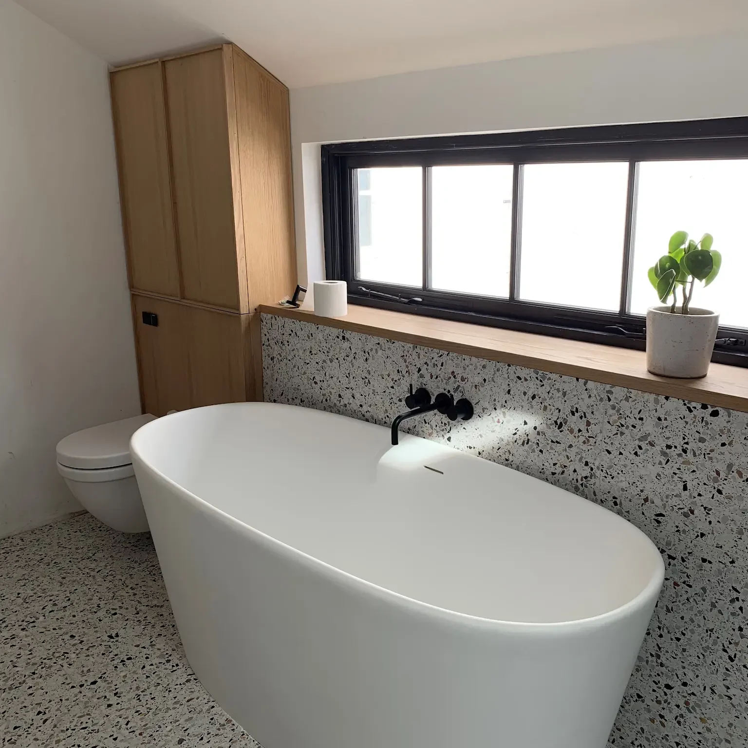 A modern bathroom featuring a white freestanding bathtub, a wall-mounted toilet, and a terrazzo tile floor. The wall behind the bath has terrazzo tiles, a black-framed window, and a small potted plant. Adjacent is a wooden storage cabinet.