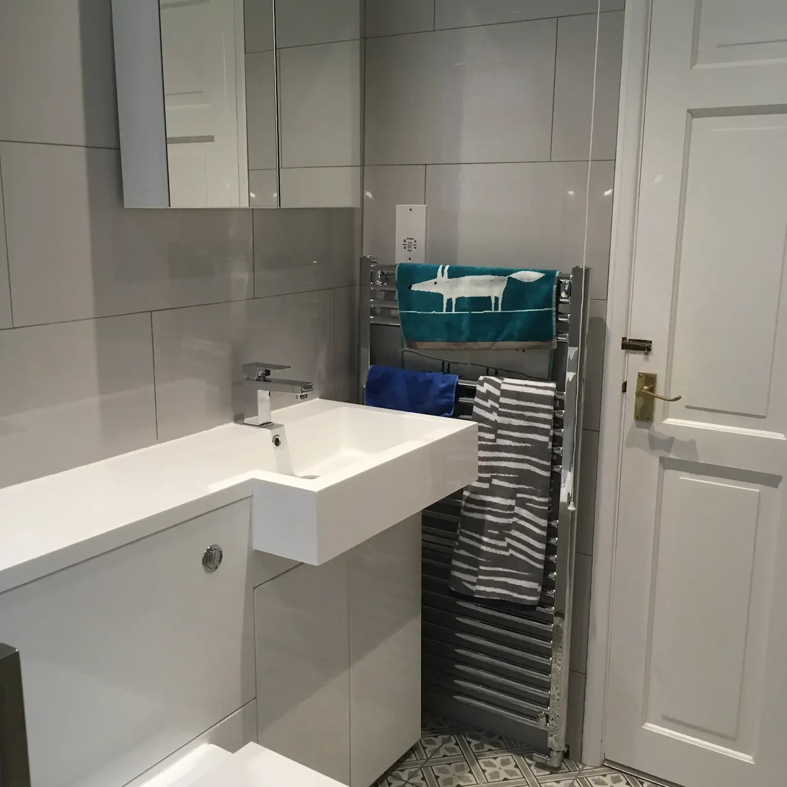 A small, modern bathroom with a white sink and a toilet under a large mirror. A white door with a brass handle is visible on the right. The gray-tiled walls and geometric-patterned floor contrast with colorful towels on a stainless steel towel rack.