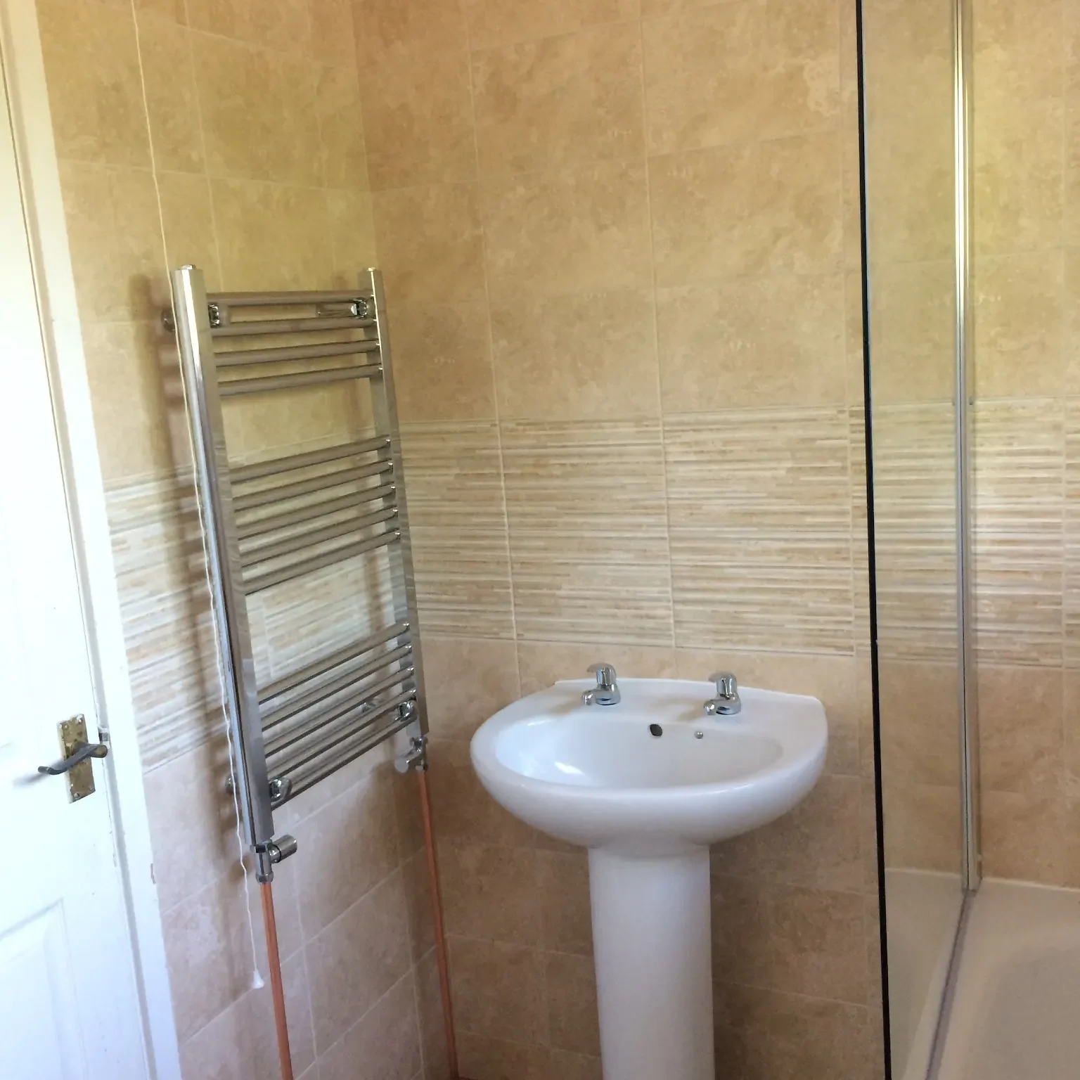 A bathroom with beige tiled walls and a white pedestal sink. Above the sink are two faucets. To the left of the sink is a towel radiator. Part of the bathtub and a glass shower screen are visible on the right. A white door is partially seen to the left.