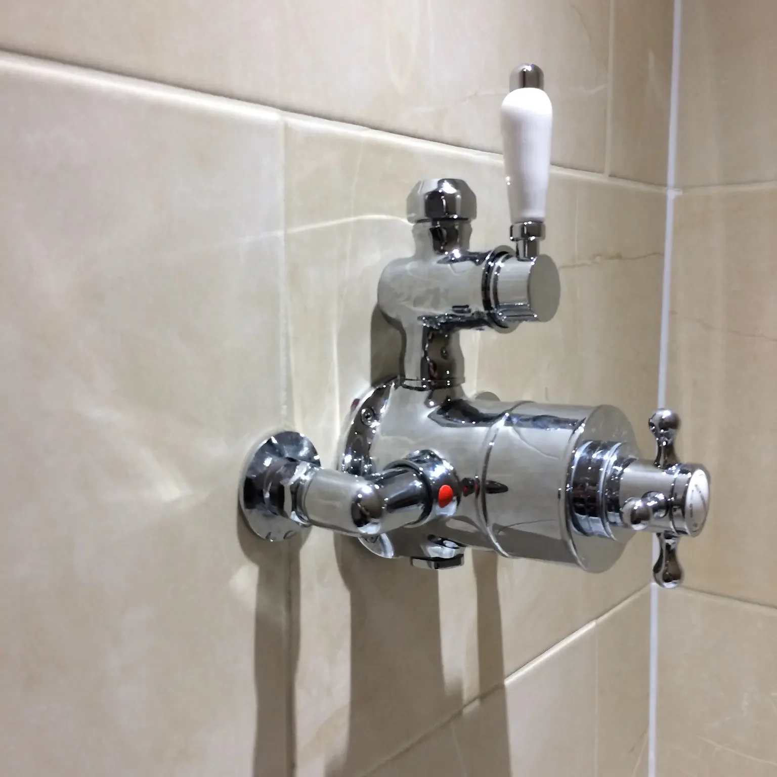 A shiny chrome shower mixer tap mounted on a beige tiled bathroom wall. The tap has two white-handled levers for adjusting water flow and temperature, and a red marker indicating the hot water setting.