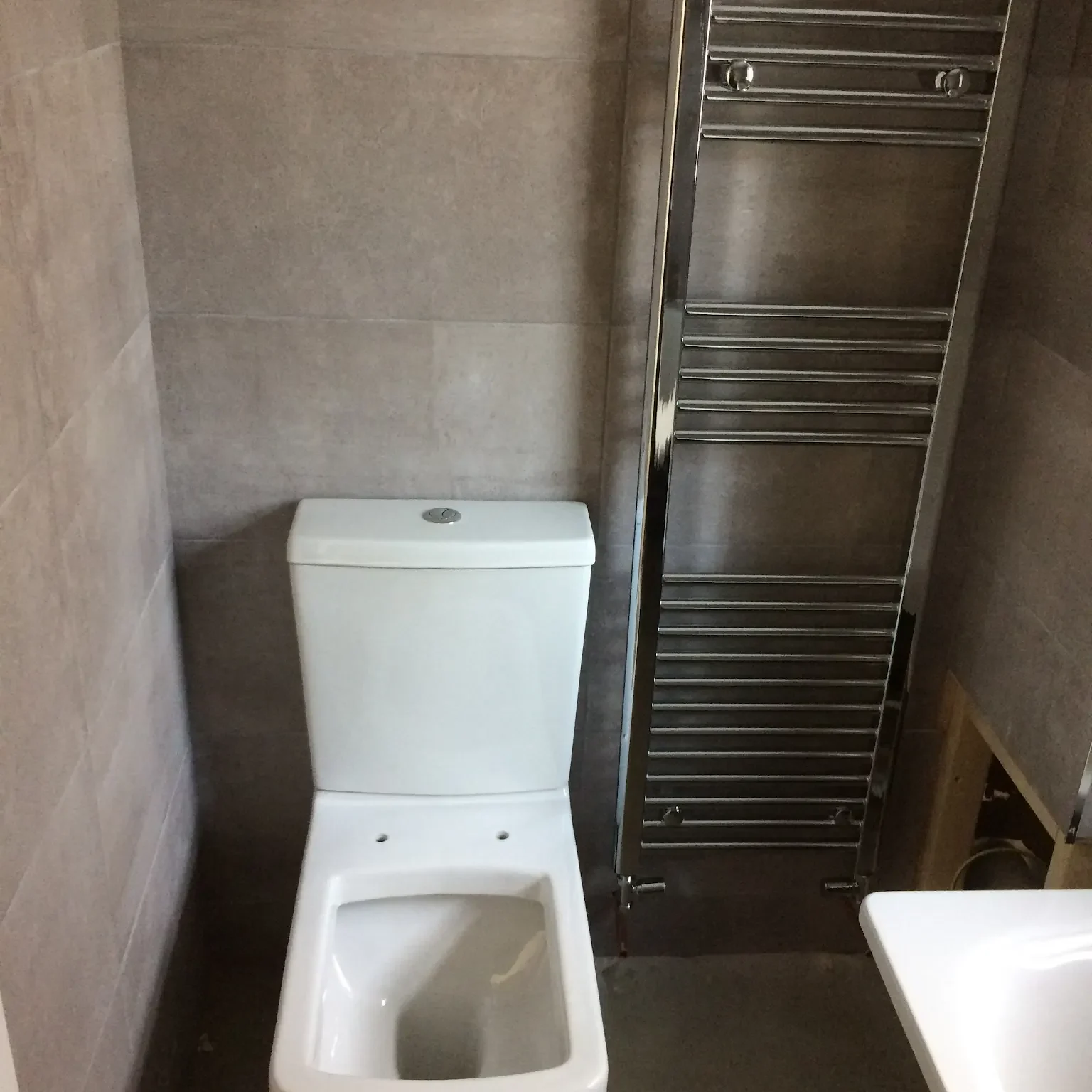 A compact bathroom features a white toilet next to a tall, sleek chrome towel radiator on the right. The walls are covered with grey tiles, creating a modern, minimalistic look. The corner of a white sink is visible on the lower right side of the image.