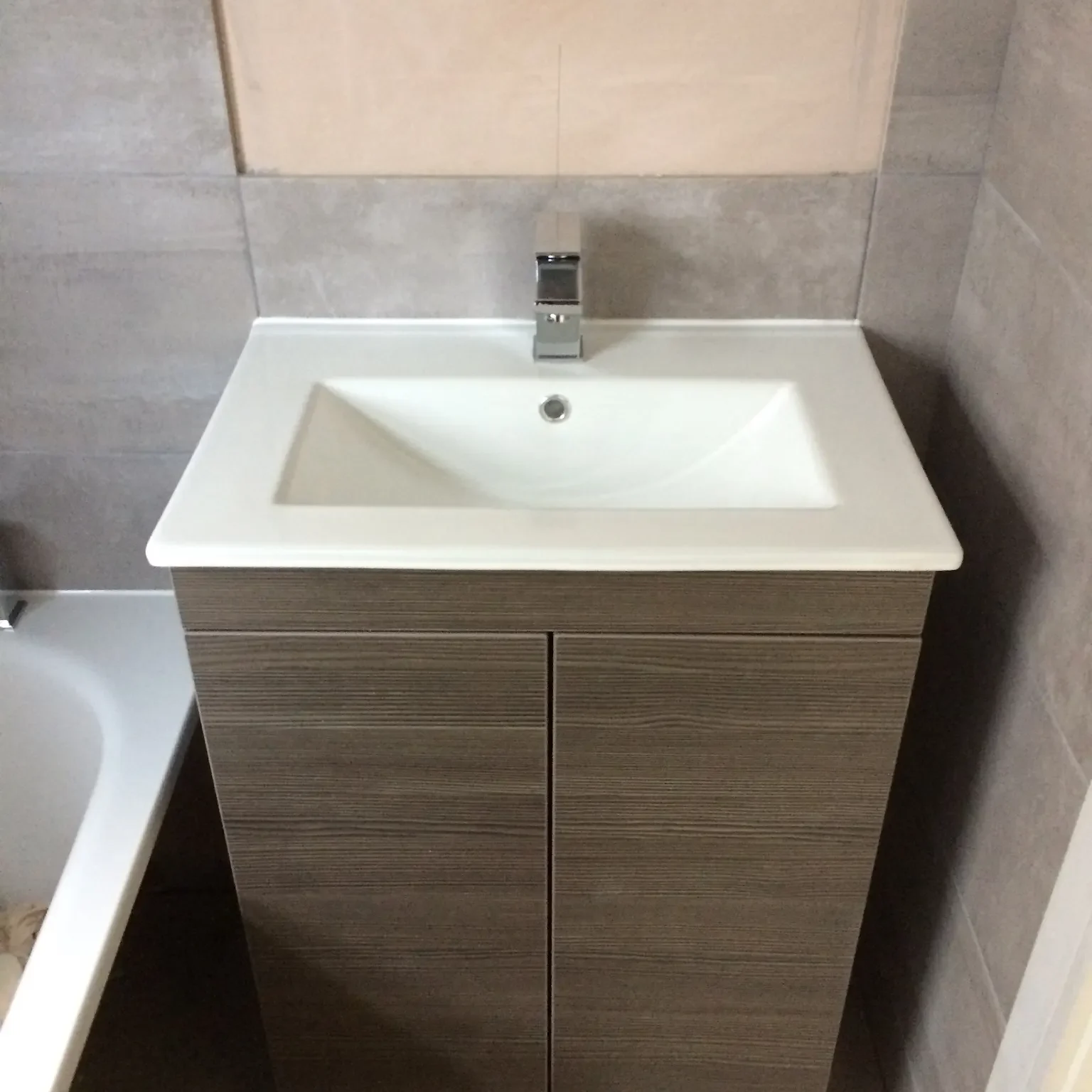 A modern bathroom vanity with a white rectangular sink and a sleek chrome faucet. The vanity has wood-paneled cabinet doors beneath the sink. The countertop is clean, and adjacent to the vanity is the edge of a bathtub. The walls are tiled in neutral tones.