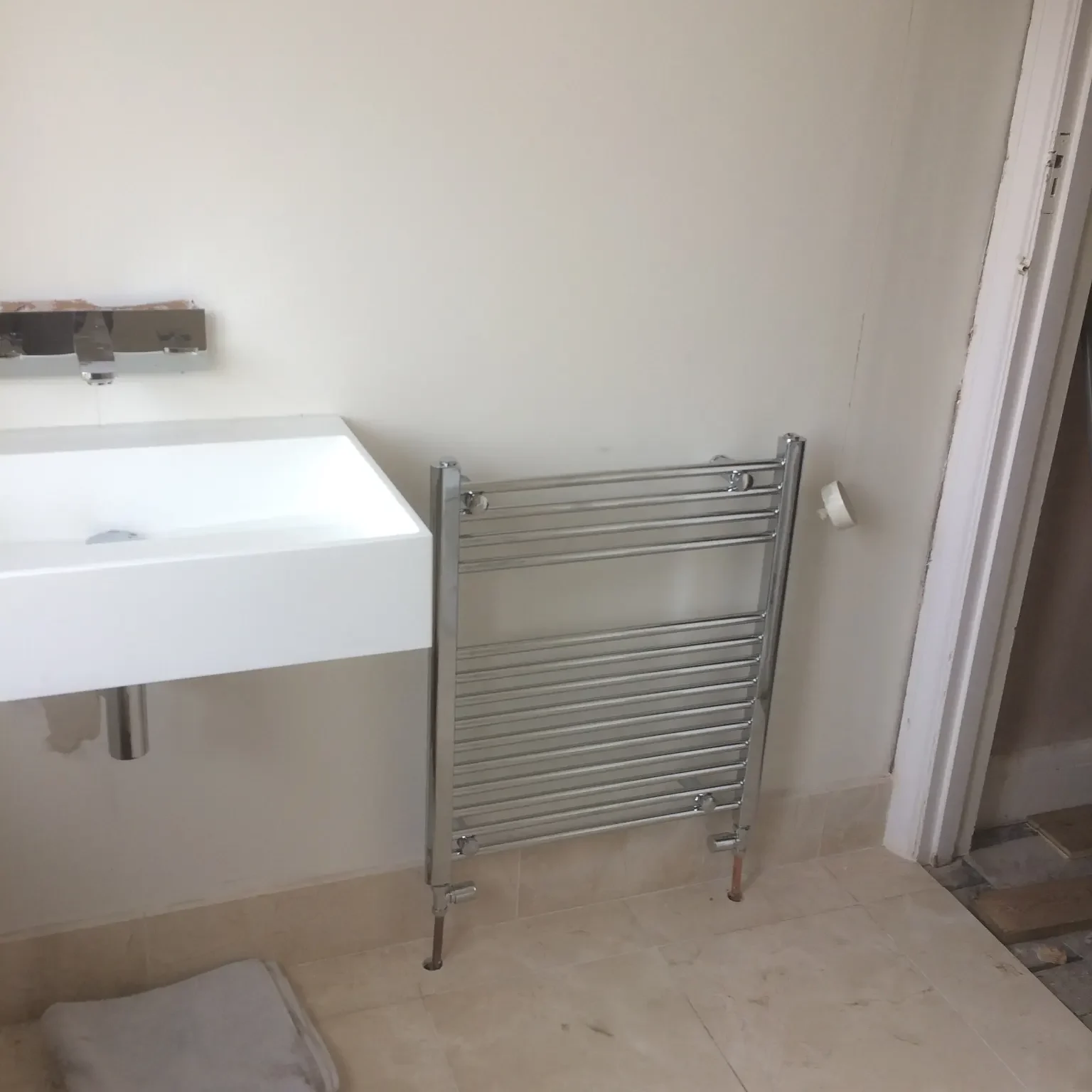 A silver towel rail is propped against a white bathroom wall beside a white rectangular sink. The floor is tiled in beige, and there is a white bath mat on the floor near the sink. A partially open white door is visible on the right side of the image.