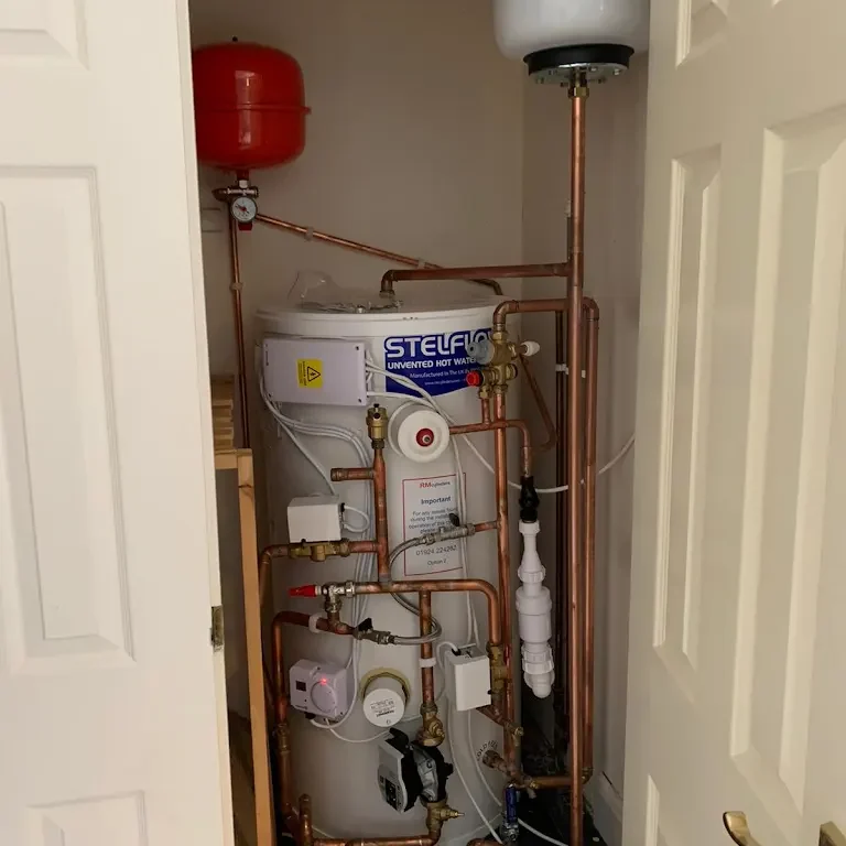 A small utility closet contains a Stelflow unvented hot water system with multiple copper pipes and fittings. A red expansion vessel is mounted on the wall above the system. The closet has double doors, one of which is partially open.