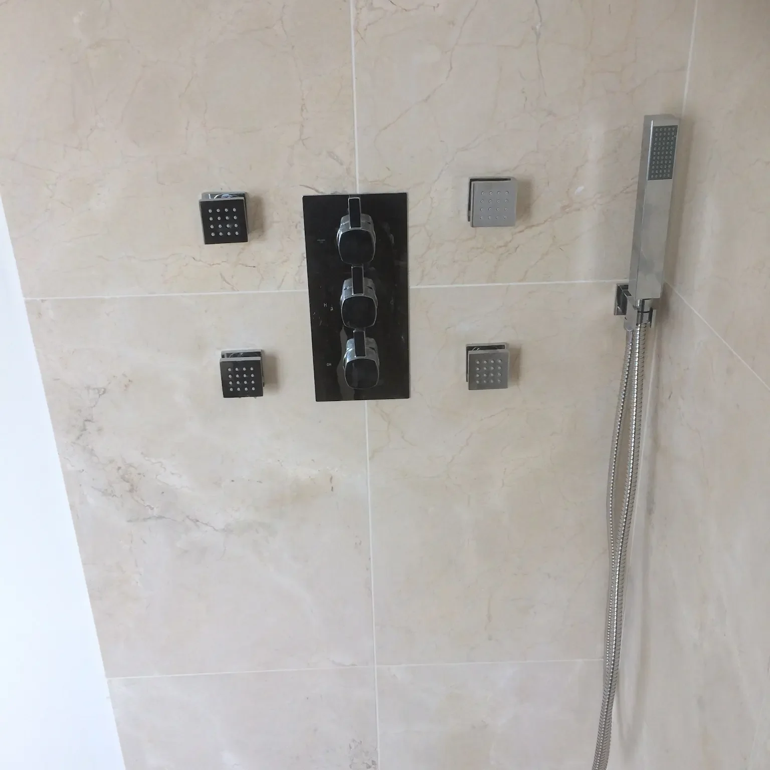 A modern shower system with a handheld showerhead attached to a flexible hose is mounted on beige marble tiles. The system includes a black panel with three circular controls and three additional square buttons arranged around it.