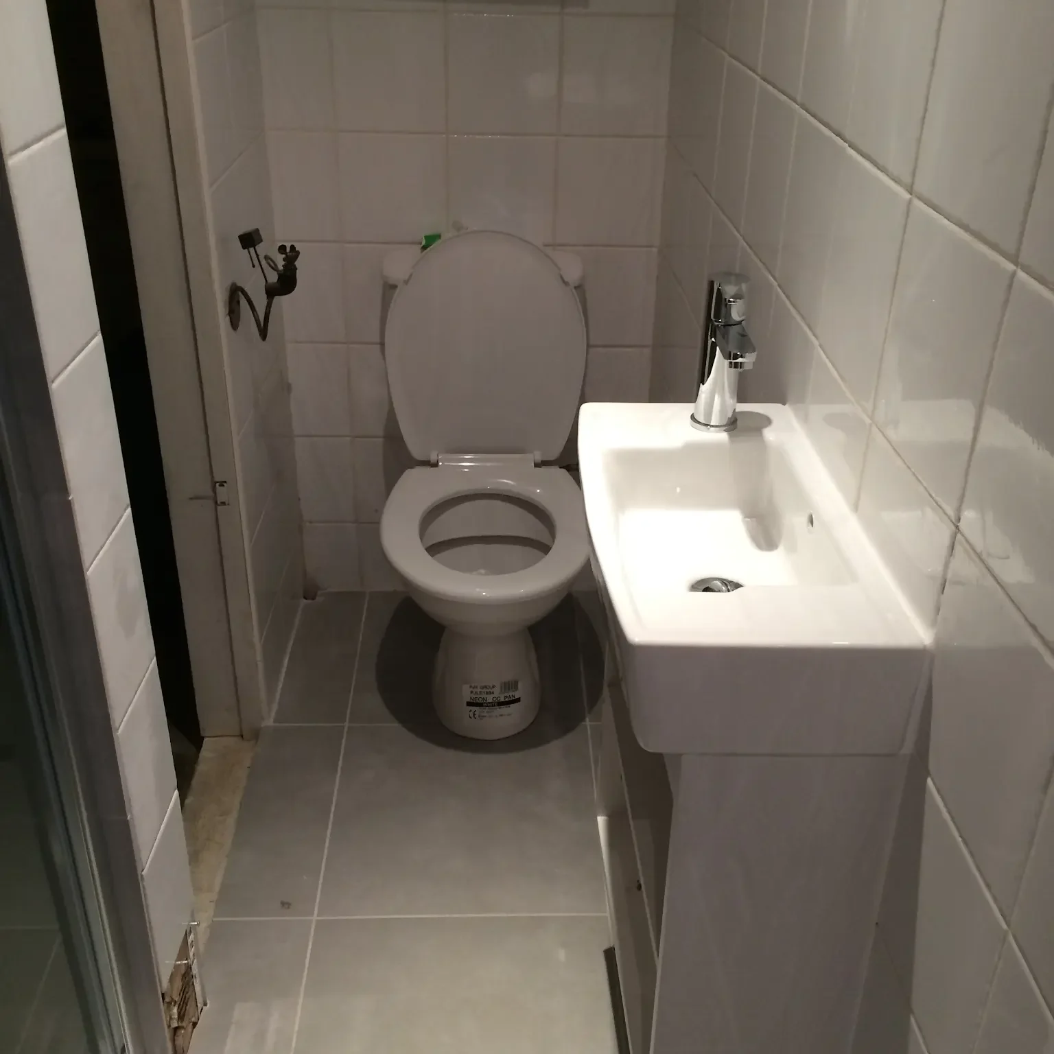 A small, narrow bathroom with white tiled walls and floor. There's a white toilet with the lid up on the left and a pedestal sink with a faucet on the right. The space appears clean and minimalist.