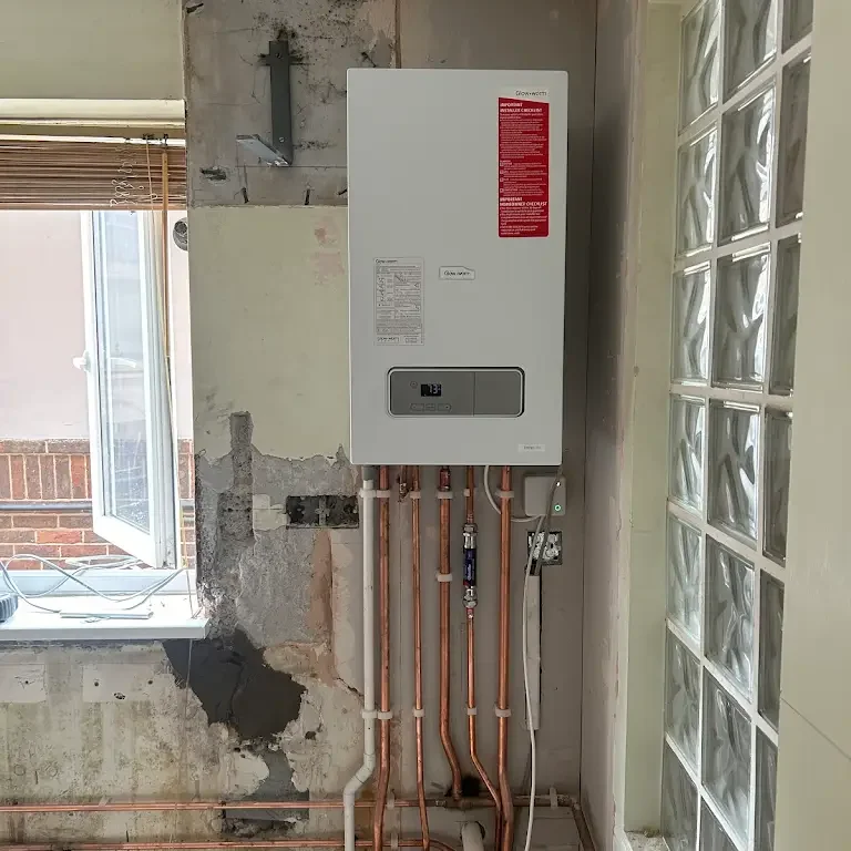 An indoor utility room with a mounted boiler system on a partially plastered wall. Copper pipes extend from the boiler, leading downward and sideways. An adjacent window with partially open blinds allows sunlight in, illuminating the space.