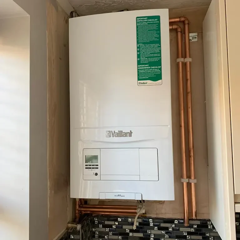 An installed white Vaillant boiler is mounted on a wall, connected with several copper pipes leading into and from it. The boiler has a digital display and a green instruction sticker at the top. The black and white tiled backsplash is visible at the bottom.